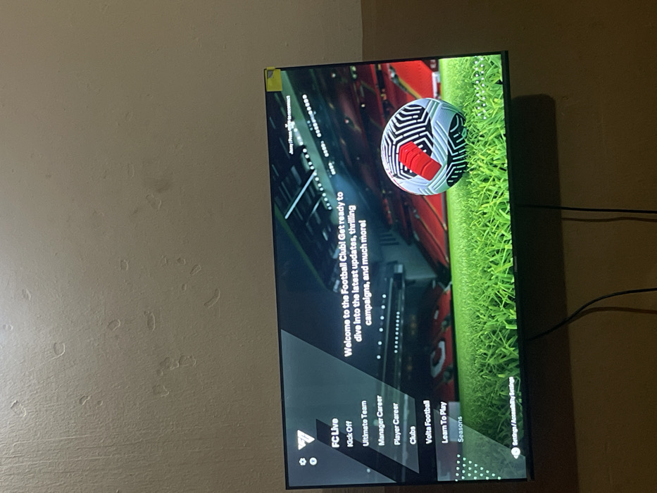 A television screen displays the main menu of a soccer game. The game options include "FC Live," "Kick Off," "Ultimate Team," "Manager Career," "Player Career," "Clubs," "Volta Football," "Learn To Play," and "Seasons." The game's tagline is: "Welcome to the Football Club! Get ready to dive into the latest updates, thrilling campaigns, and much more!" A soccer ball is visible in the background of the screen. The "Settings/Accessibility Setting" is visible at the bottom.