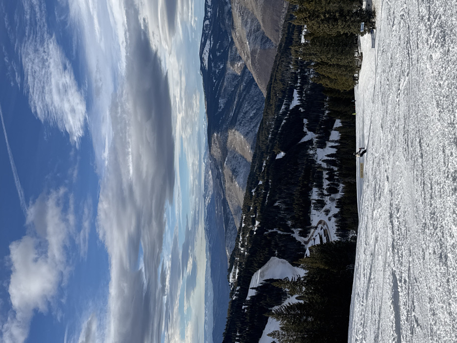 A landscape of snow-covered mountains and sky dominates the scene. The sky features blue patches interspersed with elongated white and gray clouds. Mountains stretch across the middle ground, exhibiting varying shades of brown and green interspersed with snow. A ski slope covered in textured snow fills the foreground, with a lone skier visible. Trees dot the mountainside, creating a contrast between snow and vegetation.