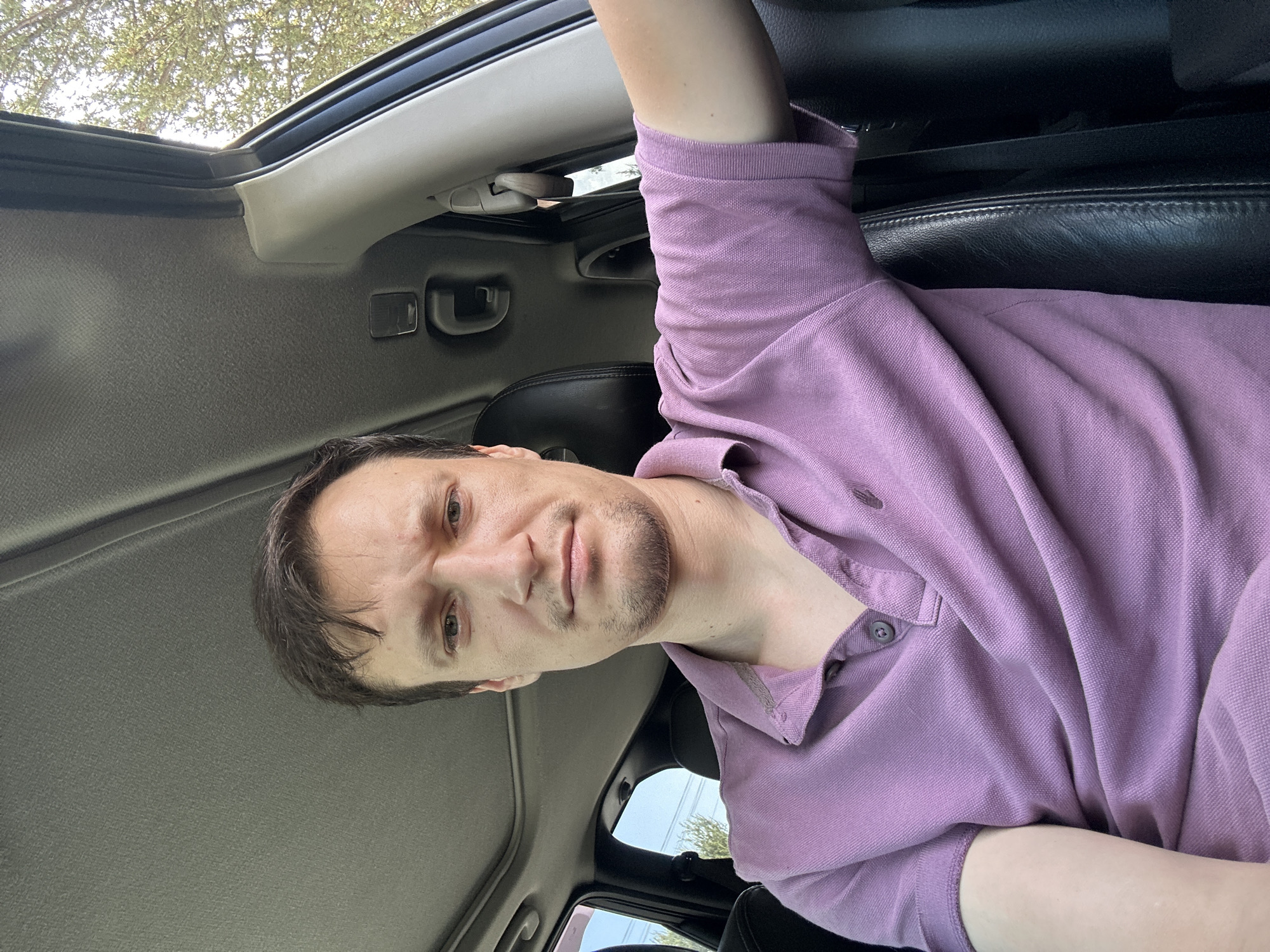 A man is sitting in the driver's seat of a car, looking at the camera. He has short brown hair and is wearing a purple polo shirt. The car's sunroof is open, and trees and the sky are visible outside the windows. The scene is lit by natural light. The location is not visible in the image.