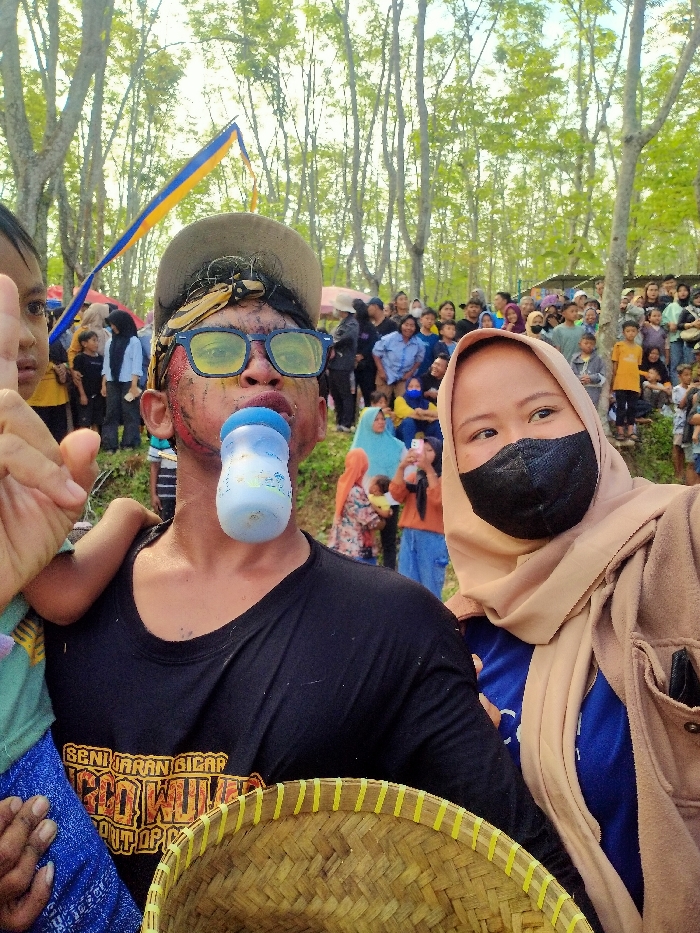 An outdoor scene in Tawangsari, Indonesia, depicts a public event with a crowd of observers. In the immediate foreground, a male individual is centered, wearing a black t-shirt with white text, a beige baseball cap, and yellow-tinted glasses. His face shows red and yellow markings, and he holds a blue and white baby bottle in his mouth. To his right, a female individual is partially visible, wearing a beige hijab, a black face mask, and a light brown jacket over a blue shirt. She looks towards the viewer's left. A child's head and arm are visible to the left of the male subject. A woven bamboo basket is partially visible in the bottom right foreground. The background features numerous tall, slender trees with light green leaves. A blue and yellow flag or streamer is suspended between trees in the upper left. A large crowd of adults and children, dressed in various casual attire, stands in the mid-ground, observing the activity.