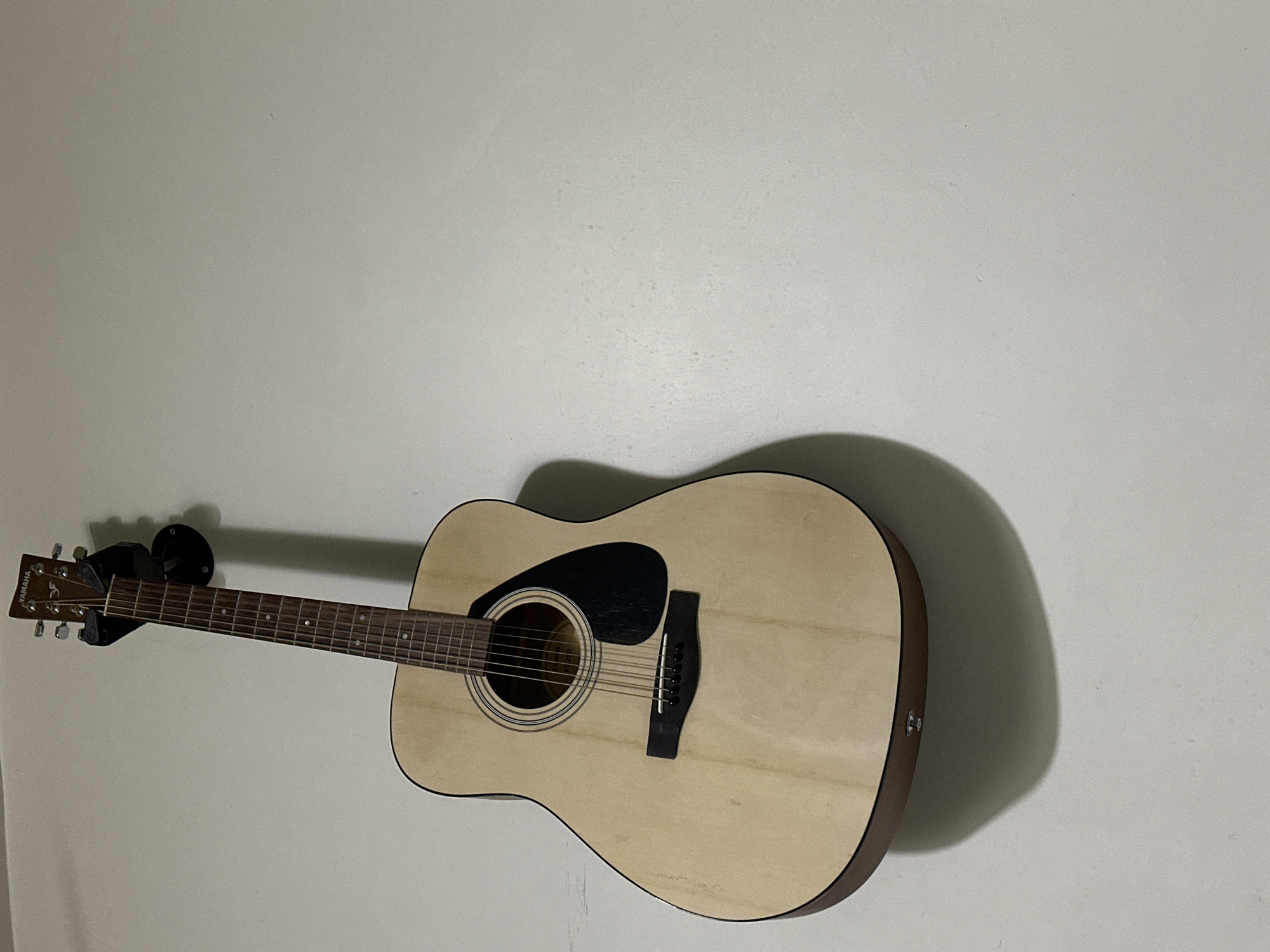 A light-colored Yamaha acoustic guitar is mounted on a wall using a black guitar holder. The wall is a light gray color. The guitar casts a shadow on the wall. The scene is indoors, under artificial lighting. No people are visible. The location is not discernable from the image.