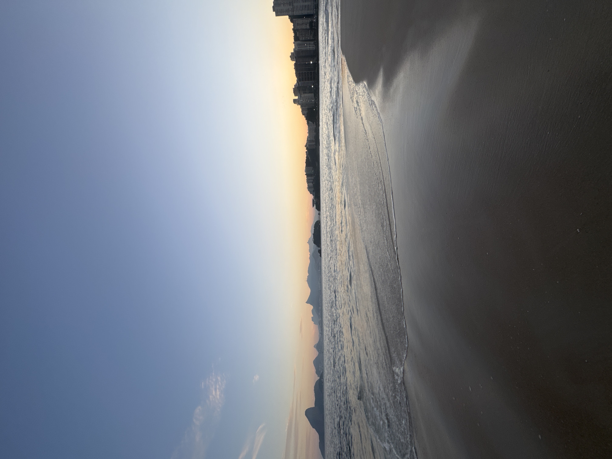 This is a photograph of a beach in Niterói, Brazil, likely during sunrise or sunset given the warm hues in the sky. The image is captured from a low angle, close to the water's edge. The foreground is dominated by wet sand, reflecting the light and showing subtle patterns from the receding waves. A gentle wave is breaking on the shore, creating a foamy white line.
In the mid-ground, the ocean stretches out, appearing calm with subtle ripples. Across the water, the distant coastline of Rio de Janeiro is visible, featuring a range of mountains. Most prominent is the iconic Pão de Açúcar (Sugarloaf Mountain) on the left, with other distinctive peaks behind it. The city skyline of Rio de Janeiro is also visible in the distance, with buildings interspersed with greenery on the hillsides.
The sky transitions from a clear, light blue at the top of the frame to warmer tones of orange and yellow near the horizon, indicating the presence of the sun just above or below the visible landscape. A few wispy clouds are present in the upper left portion of the sky. There are no people or animals visible in the frame. The overall mood is serene and natural.