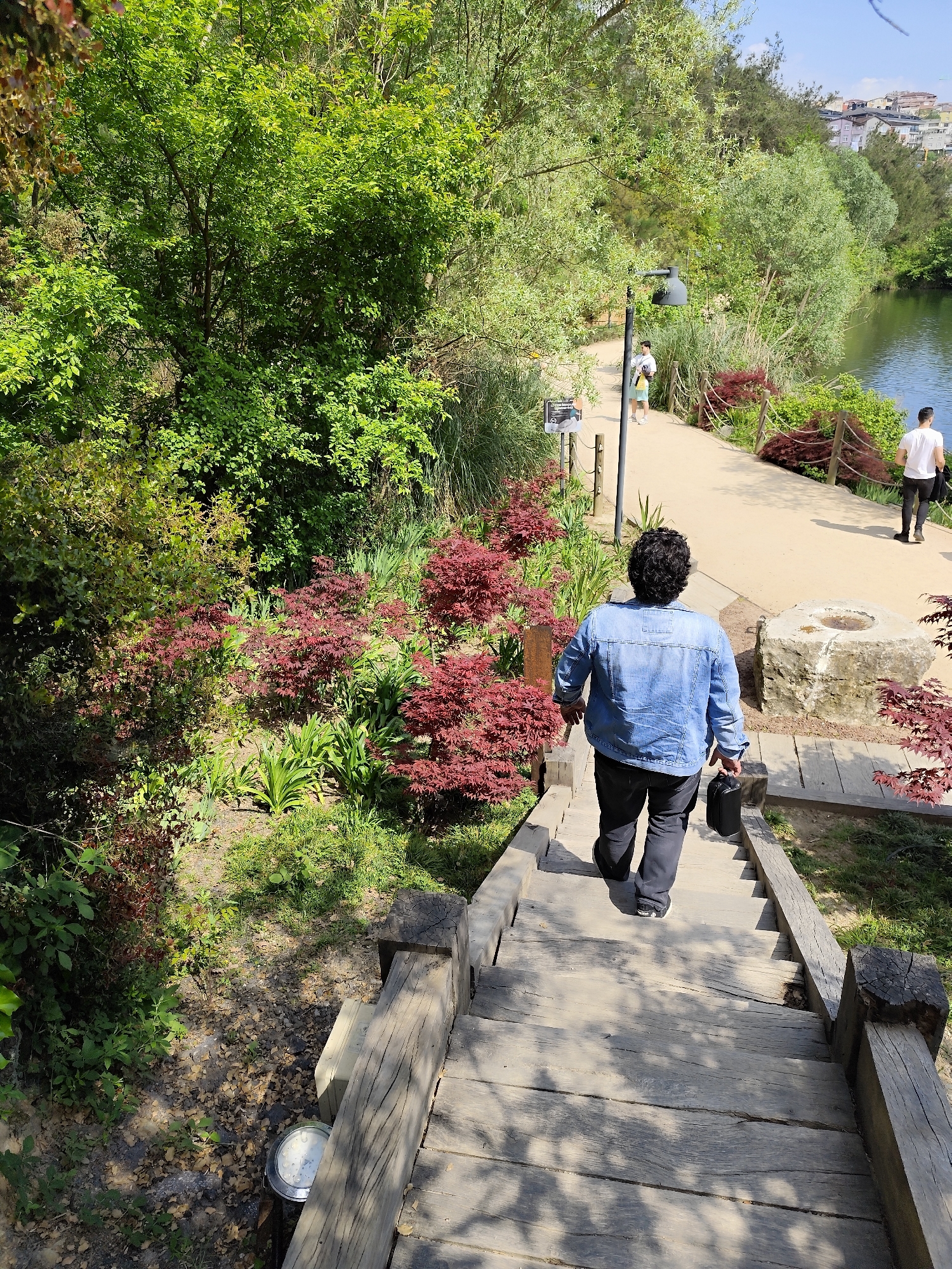 A medium shot captures a person walking down a wooden staircase in a park on a sunny day. The individual, viewed from the back, has dark curly hair and wears a light-blue denim jacket over dark pants and shoes. They are carrying a small, black, rectangular bag in their right hand.
The staircase is made of weathered wood, with railings on both sides constructed from similarly aged lumber and tree-trunk sections. The surrounding environment is lush with greenery, including mature trees, bushes, and red-leaved shrubs lining the sides of the staircase.
Beyond the stairs, a pathway is visible where two other individuals are walking. One is wearing a white t-shirt and dark pants, and the other, farther away, wears a light-colored top and darker bottoms.
The pathway runs alongside a body of water, possibly a lake, visible in the background. Beyond the water, a hillside is dotted with buildings.
Based on the landscape and architecture, this scene is located in the city of Sarıyer, Turkey.