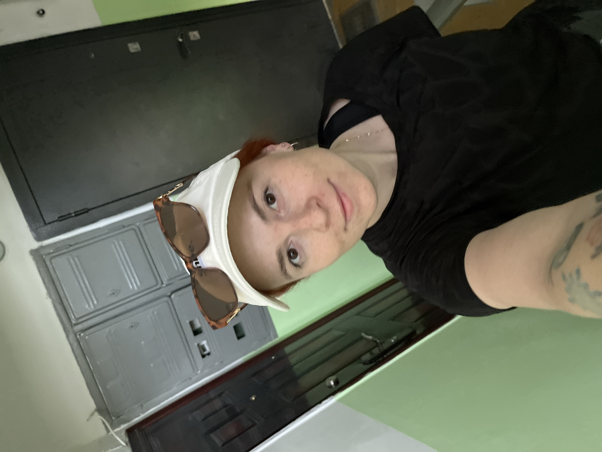 A young adult with reddish-brown hair, wearing a white baseball cap backward with tortoiseshell sunglasses resting on top, captures a selfie in what appears to be an apartment building hallway in Chaikovsky, Russia. The individual, exhibiting freckles on the face and small hoop earrings, is wearing a black short-sleeved top with shoulder cut-outs revealing a black undergarment, and a delicate gold-colored necklace. A tattoo is partially visible on the left forearm. The background features a light green painted wall on the left, transitioning to a light grey wall behind the subject. Two dark, likely metal, entrance doors are visible; one partially on the left and another on the right. Between these doors, centered on the grey wall, are two grey rectangular utility or electrical panels with multiple compartments. A dark mat is visible on the floor in the bottom right corner, suggesting an entryway.