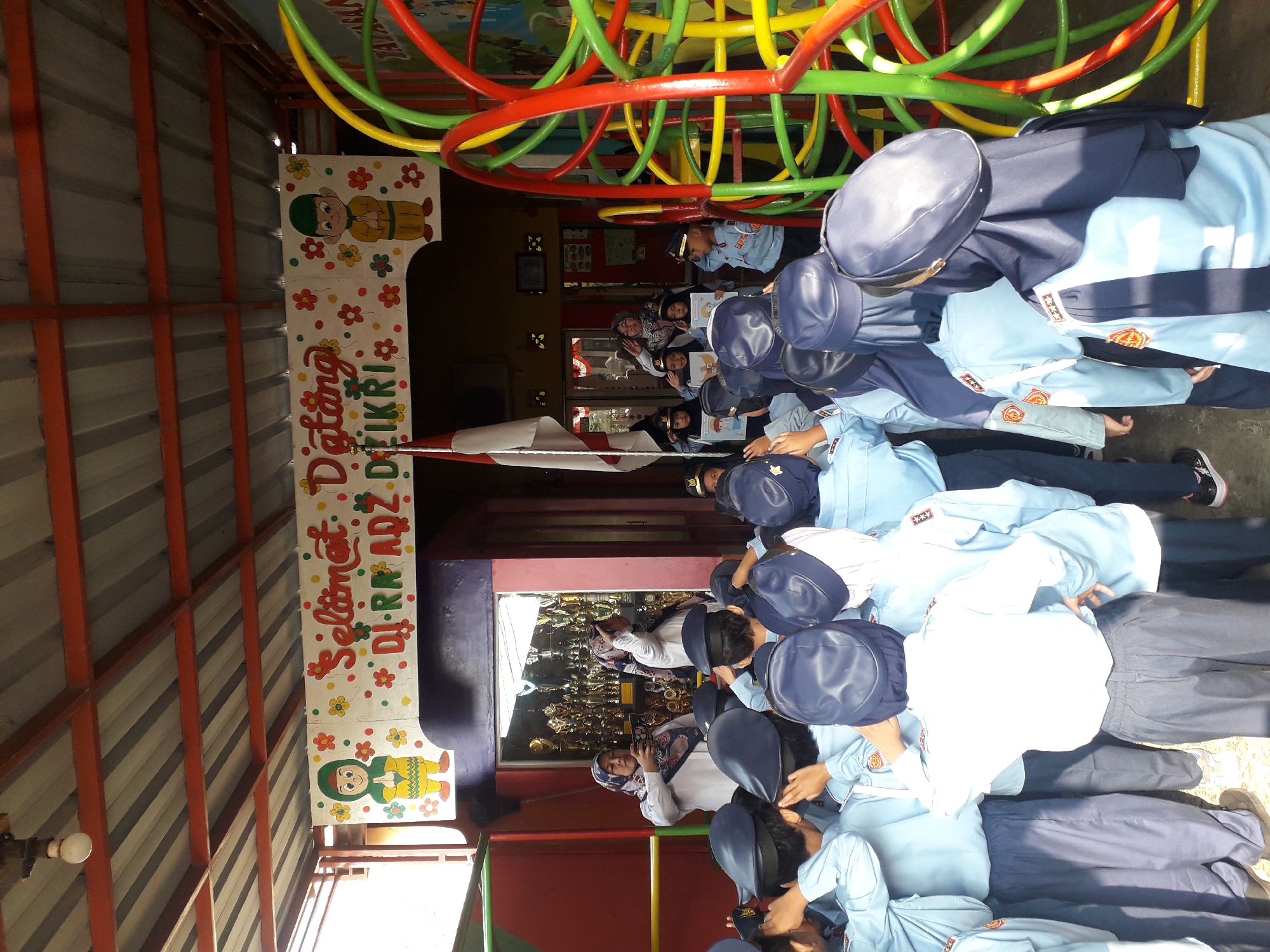 In Cankuang, Indonesia, a group of children in light blue uniforms and dark blue caps stand at attention, saluting. They appear to be participating in an event at a school or similar educational institution, as suggested by the "Selamat Datang" (Welcome) banner overhead, decorated with colorful flowers and cartoon figures. The banner hangs from a metal-framed roof structure, providing shade. Behind the children, two women in headscarves are visible; one is holding a phone, possibly taking a photo, while the other also appears to be observing the proceedings. In the background, a display case filled with trophies suggests an award ceremony or recognition event. To the right of the scene, a large, colorful playground structure with red, yellow, and green tubes is partially visible, indicating an outdoor recreational area. The overall atmosphere seems festive and celebratory, possibly marking the start of a new school term or a special occasion.