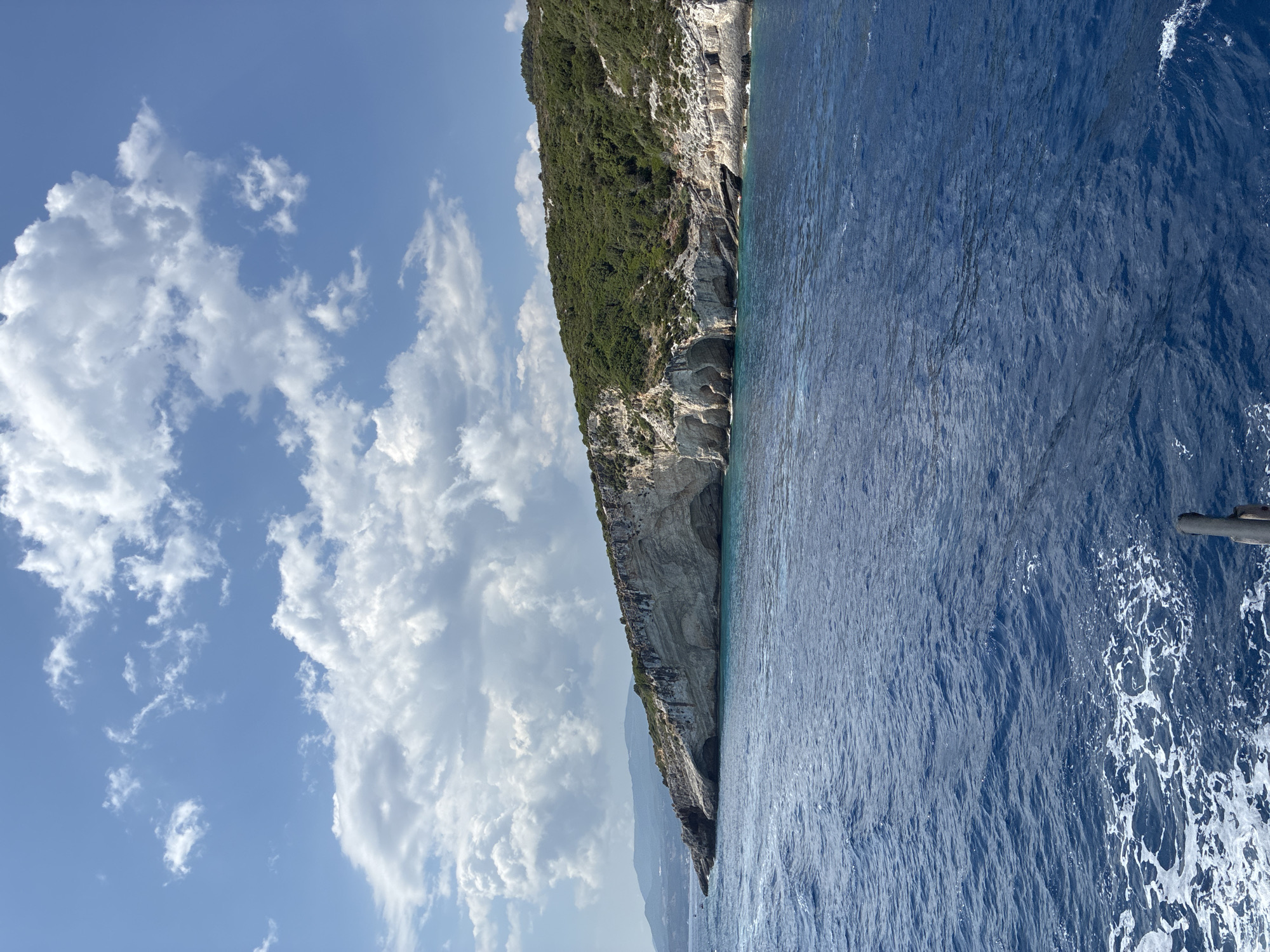 The image depicts a scenic coastal view of a rocky cliffside covered in lush green vegetation, with several caves visible at its base. The deep blue sea with white-capped waves dominates the foreground. The sky above is a clear blue, dotted with white cumulus clouds. In the distance, a hazy mountain range is visible. The scene appears to be a natural landscape, likely a popular tourist destination, possibly a sea cave area or a coastal park, located in Elaties Municipal Unit, Greece.