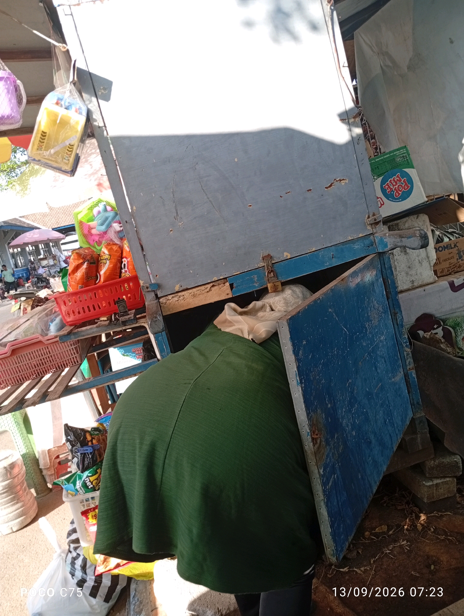 The image depicts an outdoor scene, possibly a market stall, in Jepara, Indonesia. A person wearing a green garment is partially visible, stooping over a blue wooden box with a metal latch and a padlock. The box appears to be part of a stall structure, with shelves holding various items including packaged food products, a red basket, and other goods. In the background, market activities are suggested by the presence of stalls with umbrellas and people. The ground is paved and shows signs of wear and tear. The lighting indicates it is daytime. A timestamp on the image reads "13/09/2026 07:23".