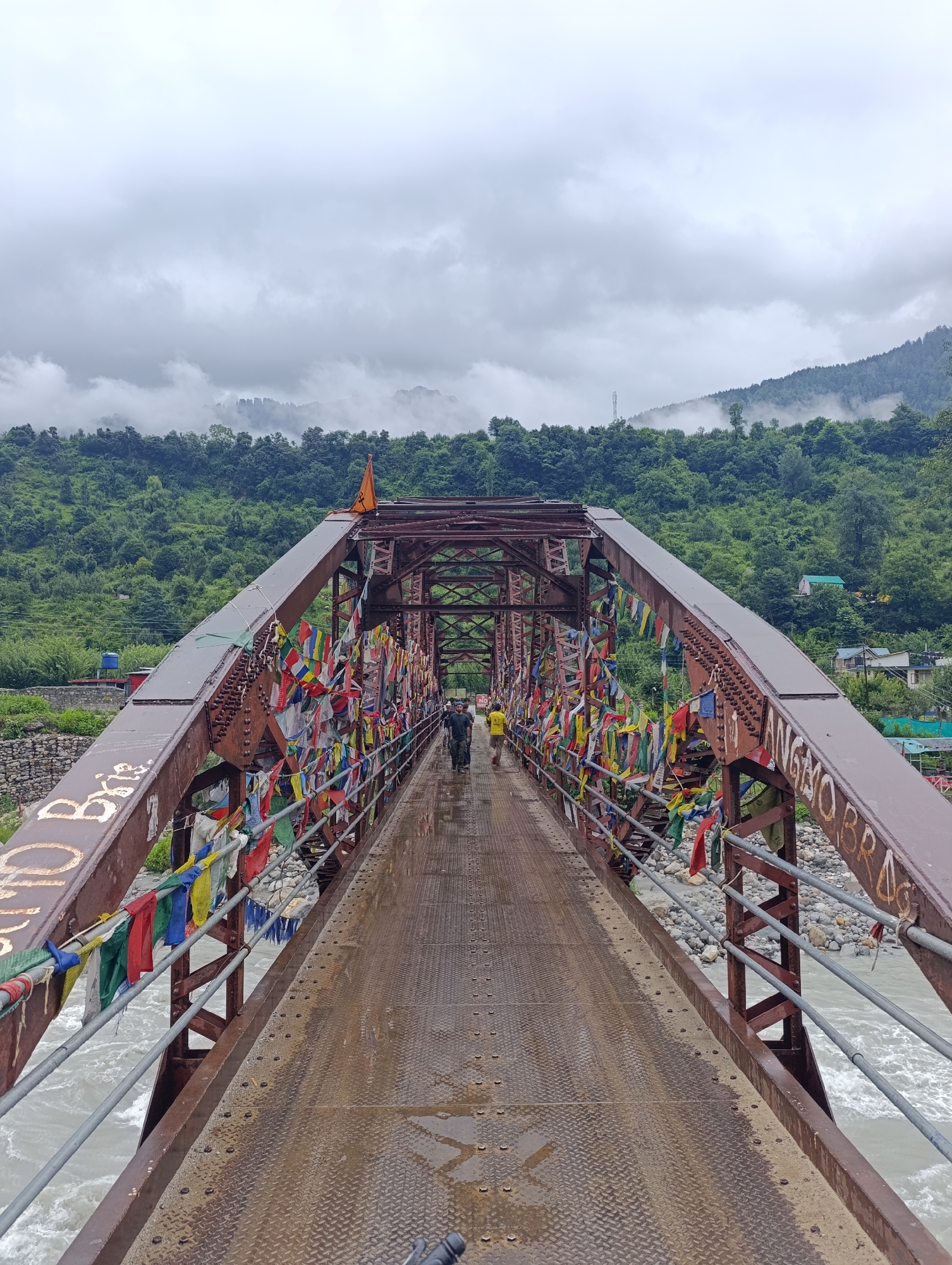 The image shows a rusty metal bridge with prayer flags strung along its railings, spanning a turbulent river. The bridge deck is wet, with visible water pooling and reflecting the overcast sky. Two people are walking across the bridge, one in darker clothing and the other in a yellow shirt. In the background, a lush green hillside covered in dense forest rises, with a few scattered buildings visible. The sky is cloudy and gray, with mist obscuring the higher parts of the mountains. The location is identified as Kalath, India.