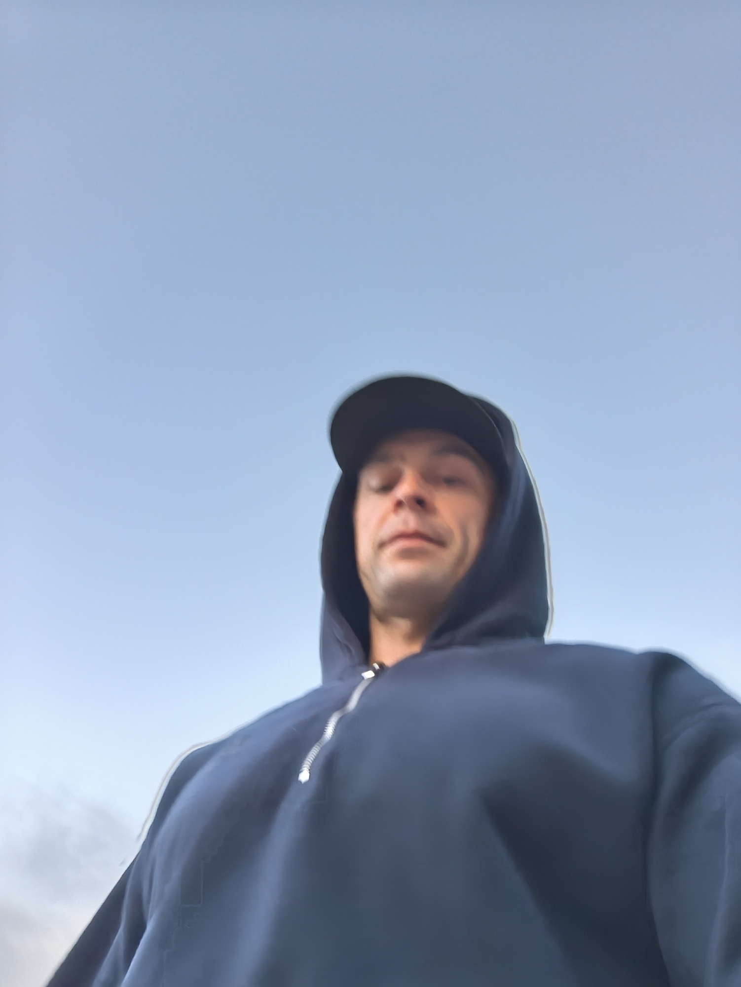A person wearing a dark blue hoodie and a black baseball cap is looking down at the camera. The background is a light blue sky. The image does not contain sufficient information to identify the location as Moscow, Russia, or to determine any specific actions or interactions.