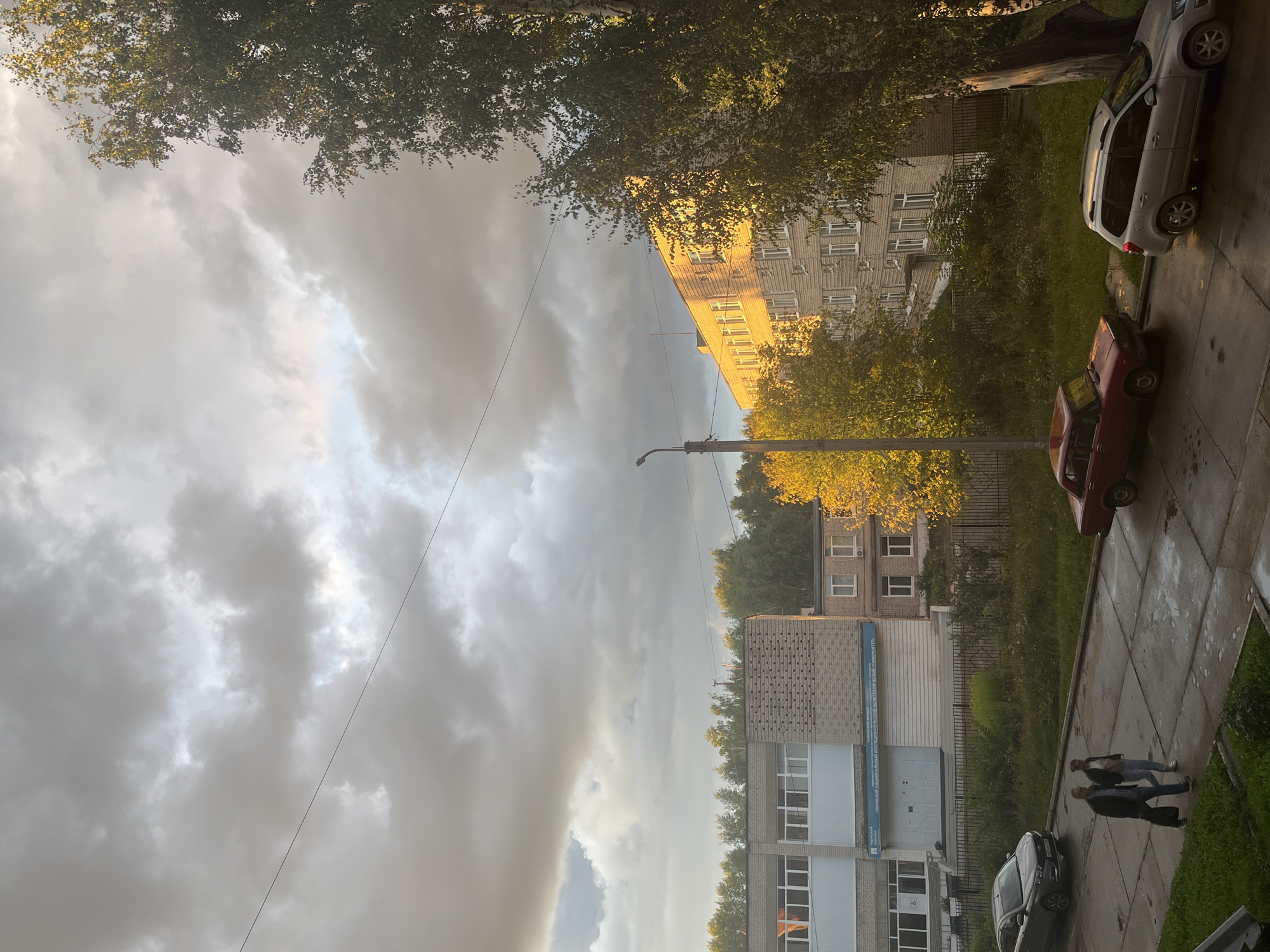 The image depicts a street scene in Chaikovsky, Russia, likely in the late afternoon or early evening, as indicated by the warm, low-angle sunlight illuminating the buildings and trees. The sky is partly cloudy with dramatic, heavy grey clouds contrasting with patches of bright, illuminated sky.
The scene features several apartment buildings, one of which is bathed in golden sunlight, highlighting its brick facade and numerous windows. In the foreground, a paved walkway runs alongside a strip of green grass and trees. Several cars are parked along the street, including a silver SUV and a red sedan. Two people are walking on the sidewalk, one slightly ahead of the other, suggesting a casual stroll.
Notable details include the presence of a street lamp and electrical wires crisscrossing the sky. The foliage on the trees appears to be turning autumn colors, with some leaves exhibiting yellow hues. The overall atmosphere is one of a slightly overcast but pleasant day in a residential urban area.
Visible text on one of the buildings appears to be in Russian. It is located on a blue banner and is difficult to read clearly due to the image resolution and angle, but it seems to be a sign for an institution or organization, possibly related to education or governance, given the common Cyrillic script and layout of such signs in Russia.