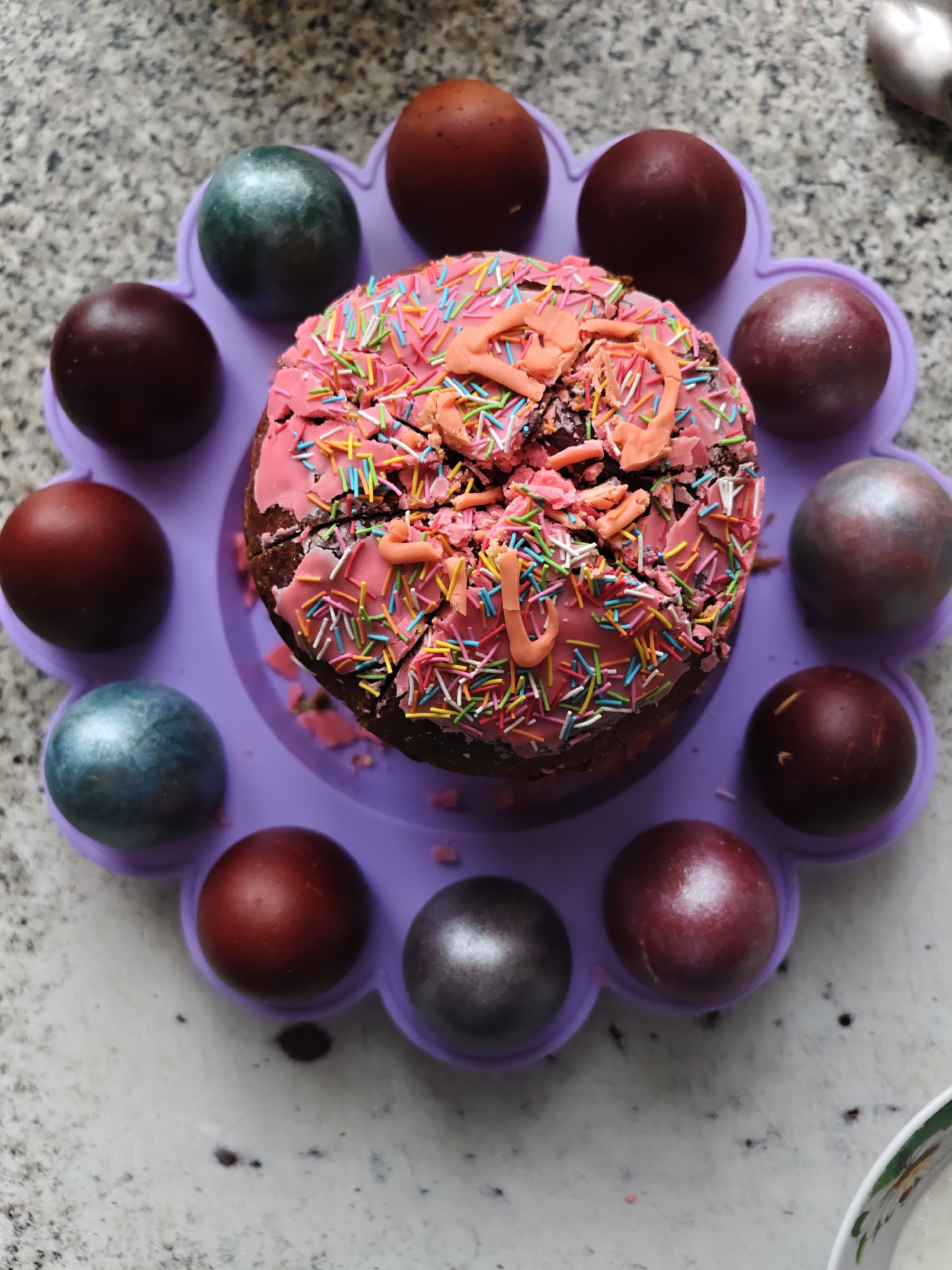 A top-down shot shows a decorated cake surrounded by dyed Easter eggs, all resting on a purple, flower-shaped silicone mold. The cake is brown and topped with cracked pink icing and colorful sprinkles, featuring a broken decoration of pink frosting on top. The eggs are dyed in shades of brown, copper, and metallic blue/silver. The background is a speckled gray surface. The image was taken in Уральское, Russia.
