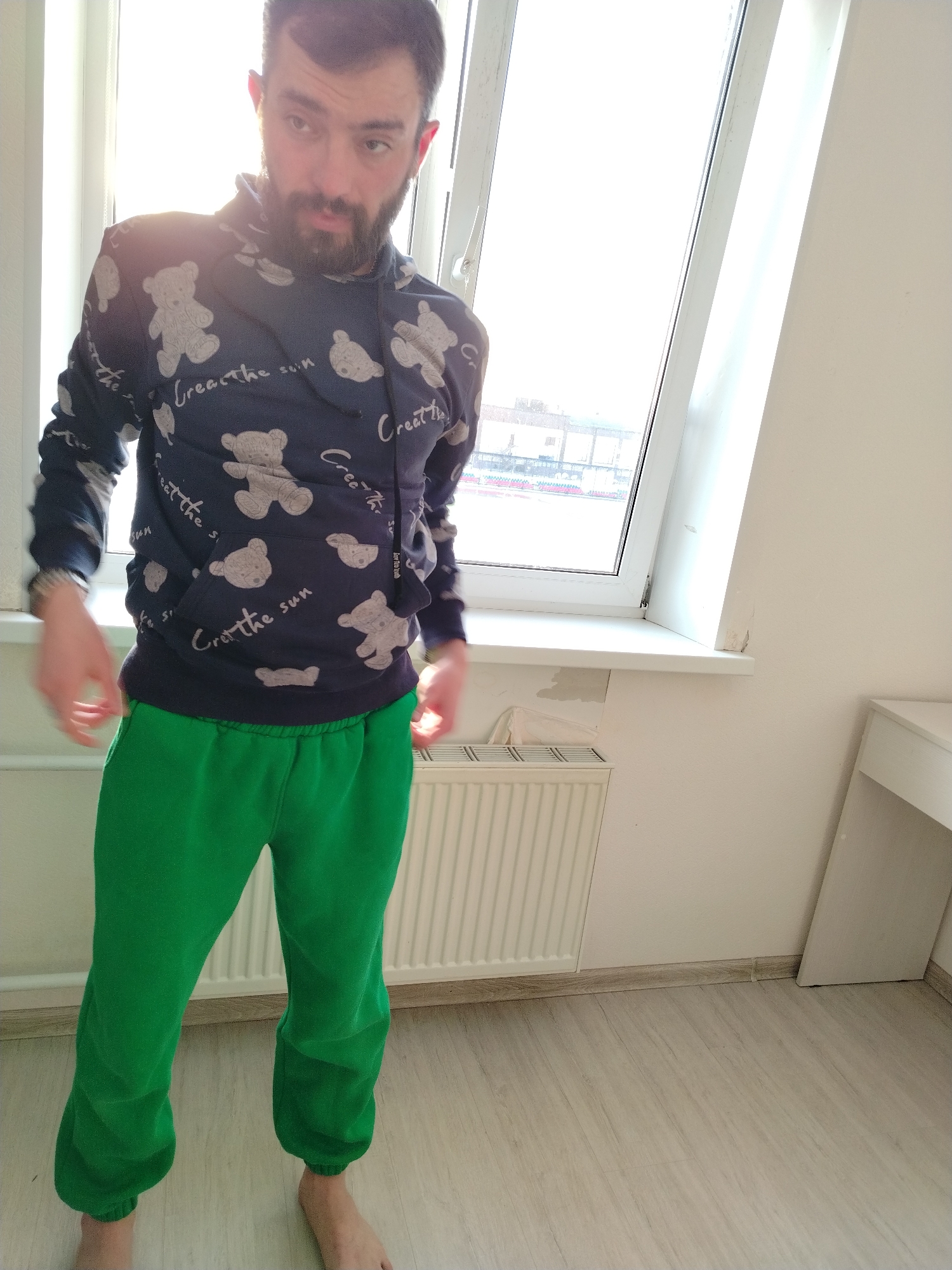 A bearded man stands in a room with light wood-look flooring. He is barefoot and wears bright green sweatpants and a dark blue hoodie printed with teddy bears and the phrase "Create the Sun." He faces the camera with a neutral expression.
Behind him is a large window with a white frame and sill. A white radiator is visible beneath the sill, and a white desk sits against the right wall. Outside the window, a building can be seen in the distance.
The scene is brightly lit by natural light from the window.
It is not possible to confirm the location as Noviy, Russia based solely on the image.