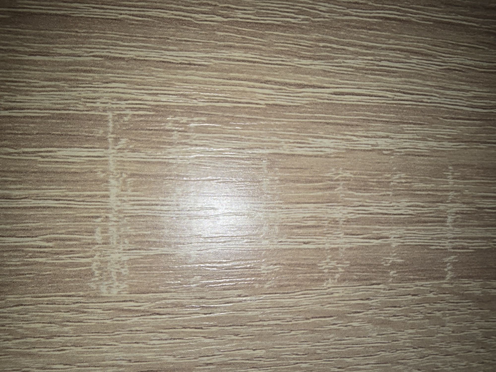 The image displays a close-up, vertical view of a textured surface resembling wood grain. The pattern consists of predominantly vertical lines of varying shades of brown and beige, with some lighter streaks suggesting the natural variations in wood. There are subtle horizontal interruptions or marks within the vertical grain pattern, particularly in the central area. The lighting appears to be from the front, creating a slight sheen and highlighting the texture. The background is entirely filled with this wood-like texture, offering no discernible context of a specific location or environment, and therefore no details about Ulan-Ude, Russia, can be provided. There are no people, subjects, objects, actions, or interactions visible in the image.