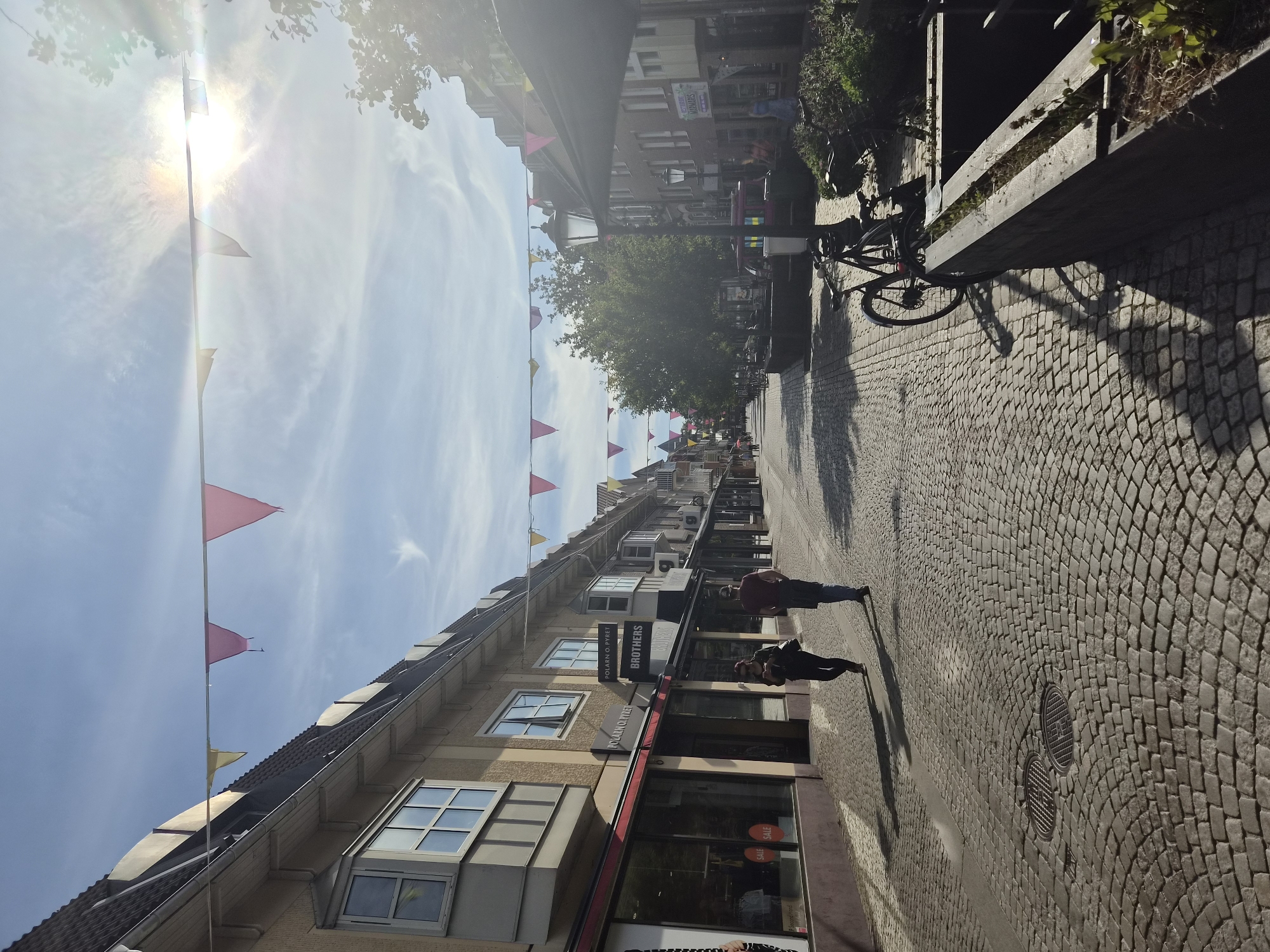 The image captures a sun-drenched cobblestone street in Varberg, Sweden, adorned with colorful triangular flags strung across the sky. The street is lined with buildings featuring shopfronts on the ground floor and windows on the upper levels, characteristic of a European town. Two individuals are walking away from the camera, their shadows stretching across the textured pavement. In the foreground, a bicycle is parked next to a planter box filled with greenery, and a streetlamp stands tall. The bright sunlight creates lens flare and casts strong shadows, indicating it's likely midday. The overall scene depicts a tranquil, pedestrian-friendly urban environment.
