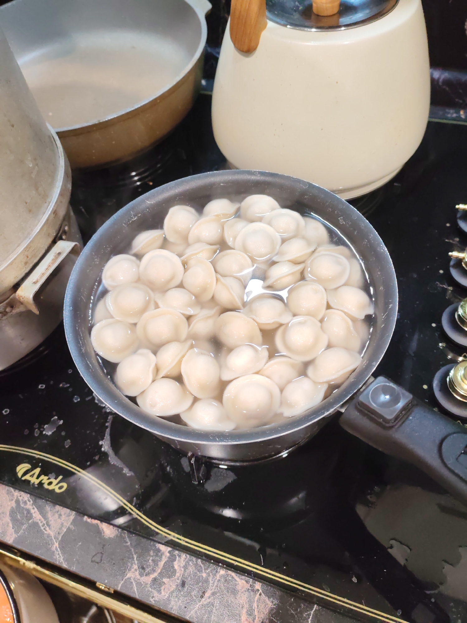 A pot filled with many small dumplings is situated on a black stovetop. To the left of the pot, a silver pot with a handle is visible. In the background, a cream-colored kettle with a wooden handle rests on the stovetop. The stovetop is clean with some water splatters around the dumpling pot. The overall scene appears to be a kitchen in a home in Тешевичи, Russia. There are no people present in the image.