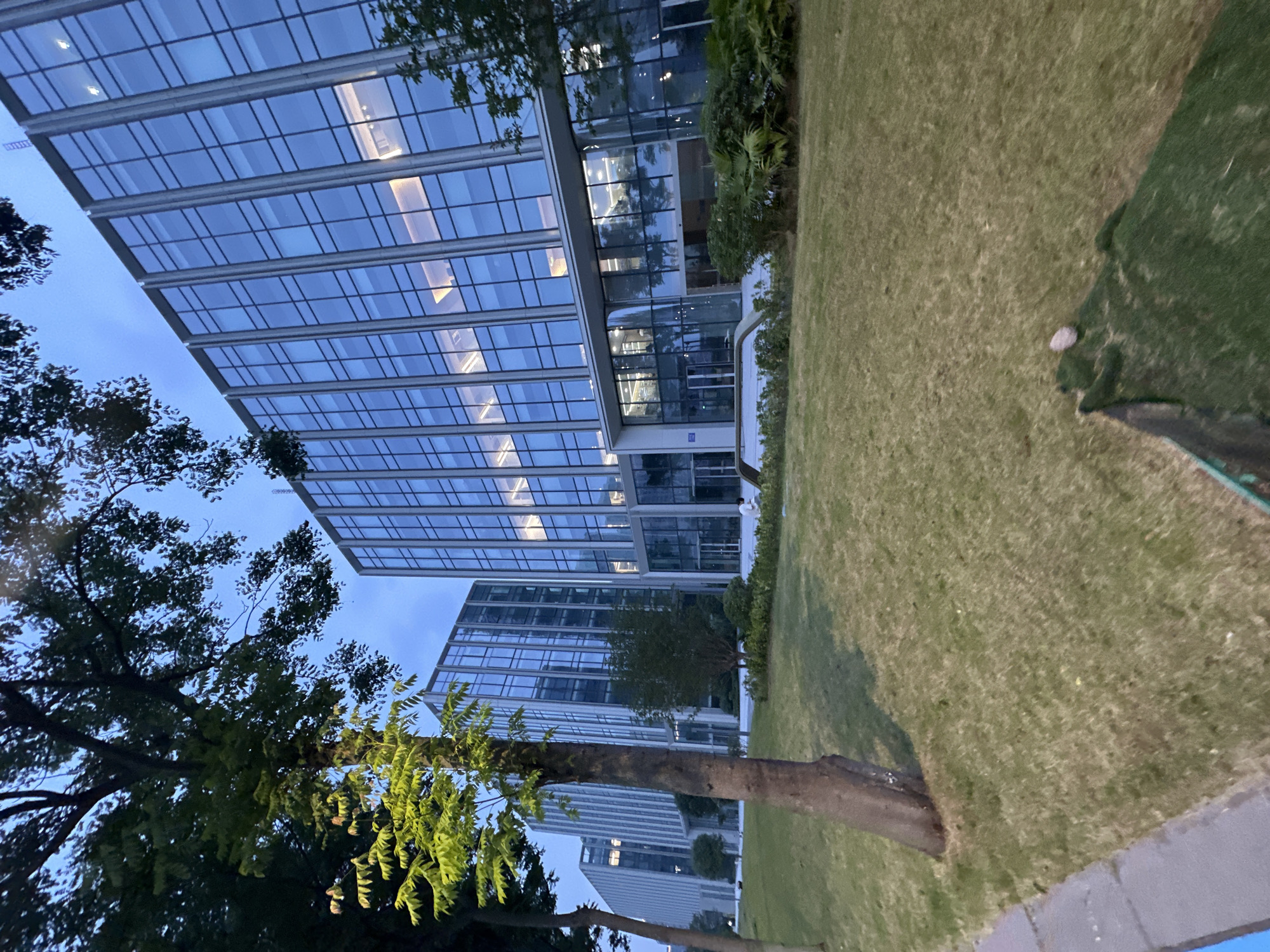 The image depicts a modern office building with a glass facade located in the Chongqing High-tech Zone, China. The building's exterior is characterized by vertical lines of windows and reflective surfaces, indicating contemporary architecture. In front of the building, a well-maintained lawn area is visible, with several trees of varying sizes scattered across the landscape. The lighting suggests it is dusk or early evening, with some interior lights on in the building, casting a warm glow through the windows. The sky is a muted blue, indicative of twilight. A single person is seated on a bench in the distance, suggesting a serene and possibly contemplative atmosphere. The overall scene conveys a sense of order and a developed urban environment.