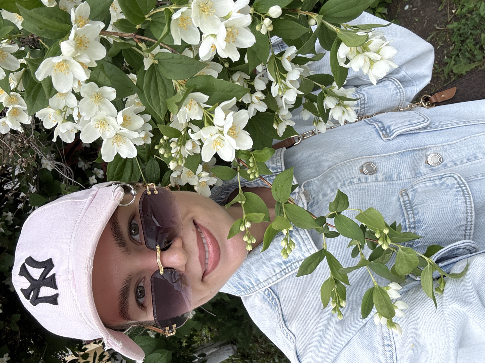 In an outdoor setting, a young woman with light brown hair and hazel eyes is captured in a selfie. She is wearing a light pink baseball cap with a black New York Yankees logo, a light blue denim jacket, and sunglasses with a gold frame. She is smiling and looking at the camera, with a branch of white jasmine flowers to her right and another branch of green leaves and white buds in the foreground to her left. The background is composed of green foliage and some dark ground. The image appears to be taken during the daytime, with soft lighting.