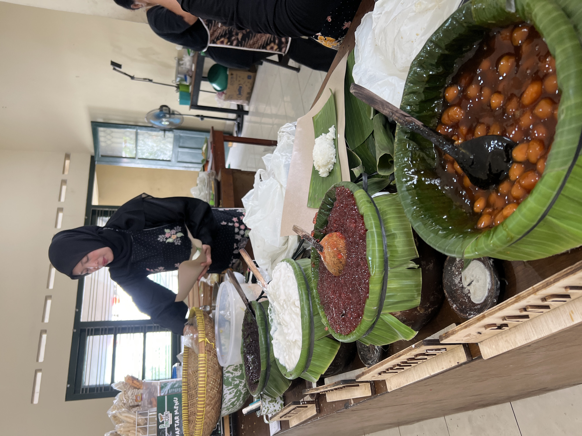 In Depok, Indonesia, a woman wearing a black hijab and an apron is serving food from a table laden with traditional Indonesian dishes. She is in the process of wrapping a portion of food in a piece of brown paper. The table displays several large bowls, some lined with green banana leaves, containing various food items. These include what appears to be black sticky rice, white rice pudding or coconut milk-based dessert, and a reddish-brown dessert, possibly made from glutinous rice, topped with an orange sauce. In the foreground, a bowl contains small, oval-shaped pieces in a brown syrup, likely a sweet dessert. There are also wooden labels in front of some of the bowls, which are common at food stalls in Indonesia. In the background, a fan is visible, and other people appear to be present, possibly customers or staff. The overall setting suggests a food stall or a small eatery.