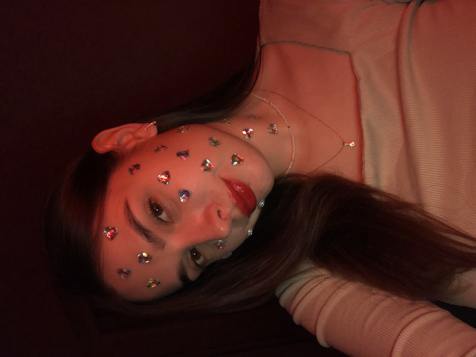 A young woman with long, dark hair is shown in a close-up, dimly lit selfie. Her face is adorned with small, heart-shaped rhinestones scattered across her forehead, cheeks, and chin. She is wearing bright red lipstick and a ribbed, light-colored top with a square neckline. A delicate beaded necklace and a pendant necklace are visible. The overall lighting is warm and reddish, casting a glow on her skin. The image does not provide any information about the city of Noviy, Russia, or any specific location.