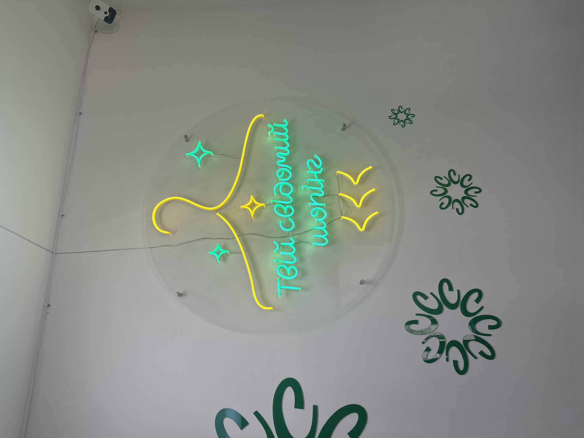 The image depicts a close-up, eye-level shot of a neon sign mounted on a white wall. The sign is circular with a yellow neon outline of a hanger at the top. Inside the hanger, the words "Твій свій одяг" are written in vibrant teal neon. Below this, in yellow neon, are three stylized chevron shapes pointing downwards. The sign is illuminated by neon lights, casting a glow on the wall. To the left of the sign, a large green stylized swirl or flourish is partially visible. Several smaller green circular embellishments resembling stylized flowers or snowflakes are scattered on the wall around the main sign. A security camera is visible in the upper right corner of the image. The lighting suggests an indoor environment. The image location is identified as Sarny, Ukraine.