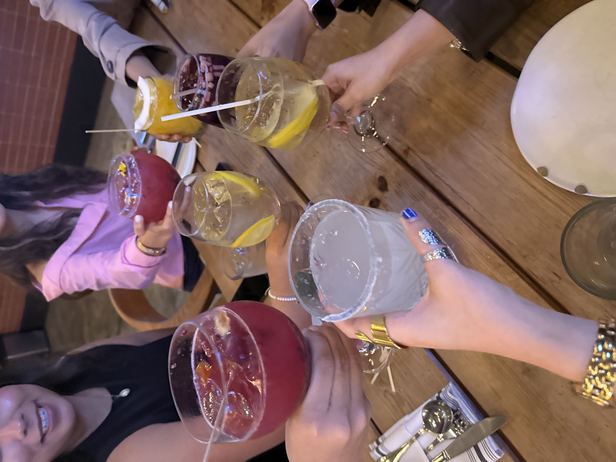 A group of people is gathered around a wooden table, raising glasses in a toast. The glasses contain various colorful beverages, likely cocktails, with ingredients such as fruit and ice visible. The table is set with cutlery and napkins. The location is likely a restaurant or bar in Mexico City, Mexico, based on the style of the drinks and the atmosphere. The people are interacting and celebrating, creating a convivial atmosphere.