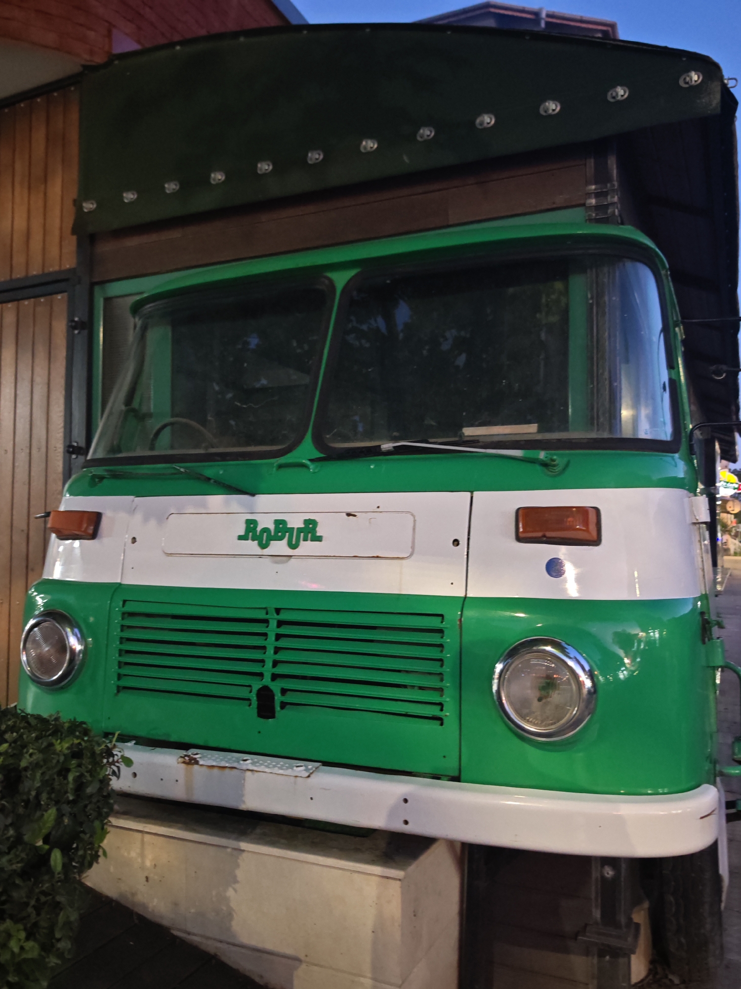 This image depicts a green and white Robur truck parked in Nesebar, Bulgaria. The truck has a green hood and grill with a white stripe across the middle, bearing the Robur logo. The windshield is large and divided into two sections. The truck bed is covered with a dark green tarp. The background shows glimpses of nearby buildings and streetlights, suggesting an urban setting. The lighting suggests the photo was taken in the evening. A bush is partially visible on the lower left corner of the image. The bumper is white and shows some signs of rust and wear. The truck is parked on a concrete base.