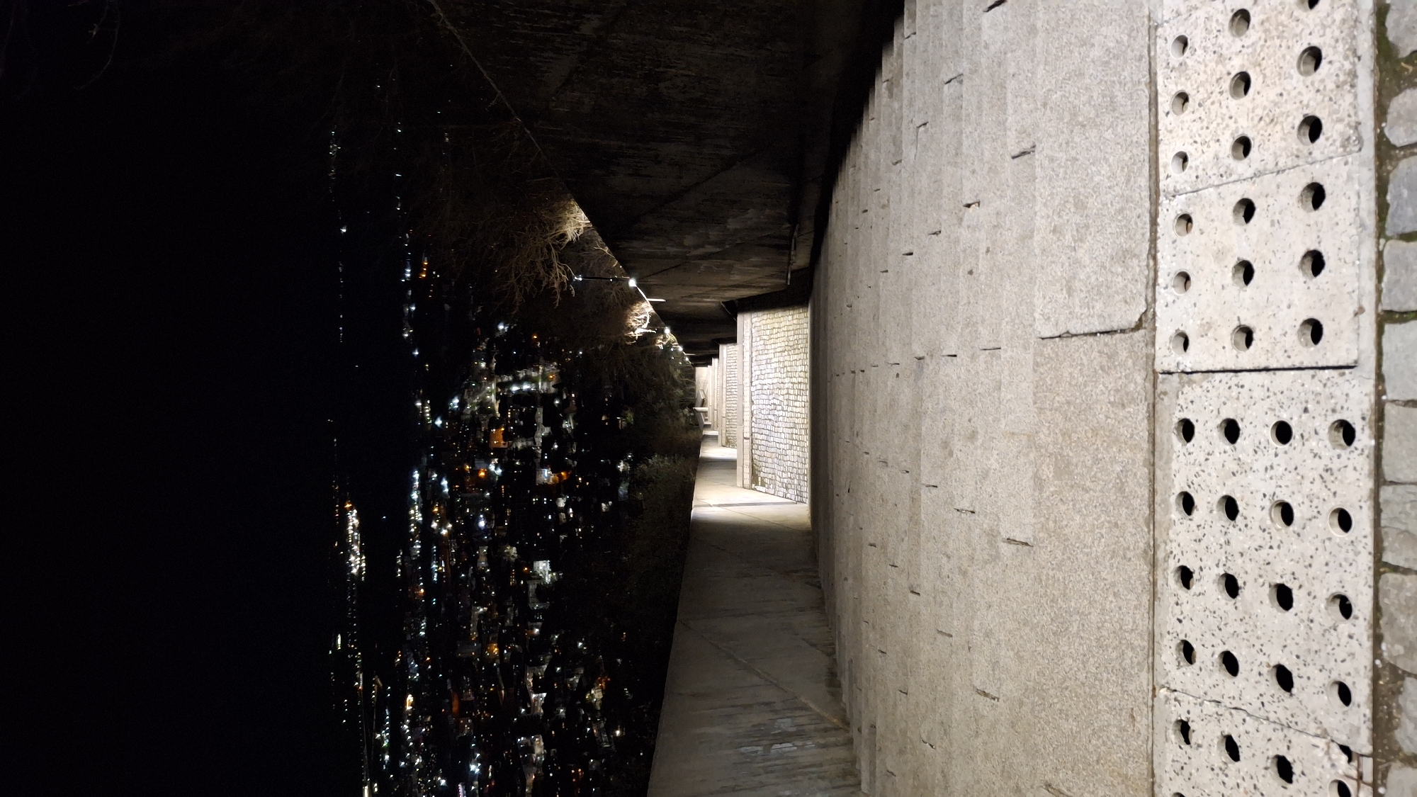 The image shows an open corridor in Shumen, Bulgaria, likely an outdoor walkway or underpass. The left side of the corridor features a dark, almost completely black area, possibly a view of the city at night with faint lights visible. The right side is a concrete wall made of large blocks, some with circular holes. The corridor extends into the distance with brick walls visible down the path. The overall setting is dimly lit and urban.