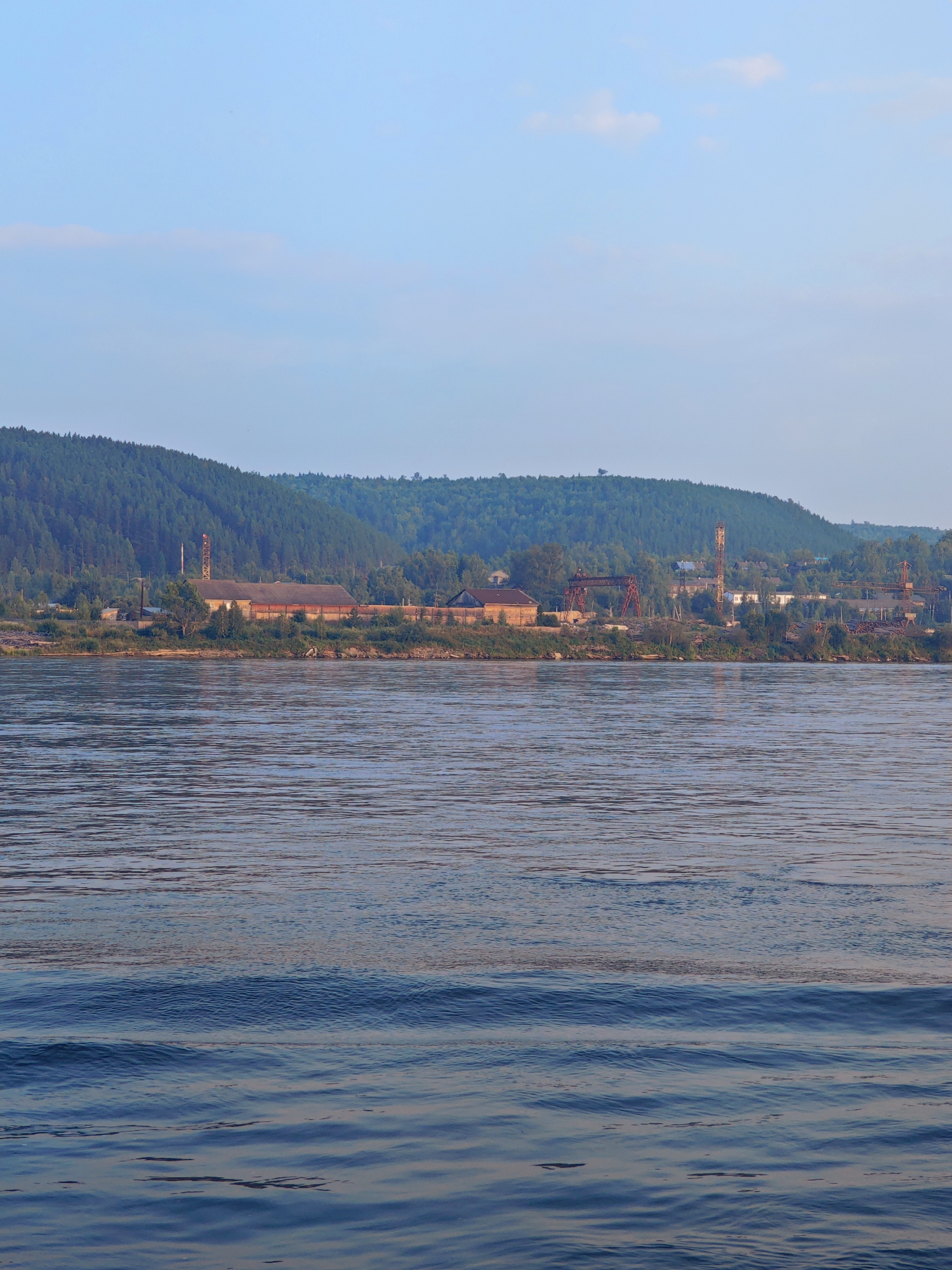 The image captures a wide river flowing in the foreground, with gentle ripples on its surface reflecting the light. In the mid-ground, a treeline of dense, dark green coniferous trees covers a sloping hill that rises behind the riverbank. A settlement is visible along the riverbank, nestled between the water and the forested hill.
The settlement appears to be industrial, with several buildings, including what looks like a long, low-slung brick structure with a corrugated roof. There are also tall, metallic structures, possibly cranes or industrial towers, with a rusted red color. Some of these structures have a lattice-like design. Scattered among these industrial elements are smaller buildings, some with brick facades and others with lighter colored walls. Smoke or steam can be seen rising from one of the taller structures. The presence of logs or timber piled near the riverbank suggests a logging or wood processing operation. The overall impression is one of an industrial area situated in a natural, forested landscape. The sky above is a clear, pale blue with a few wispy clouds. This scene is consistent with the appearance of Predivinsk, Russia, a town located on the banks of the Angara River, known for its industrial activities.