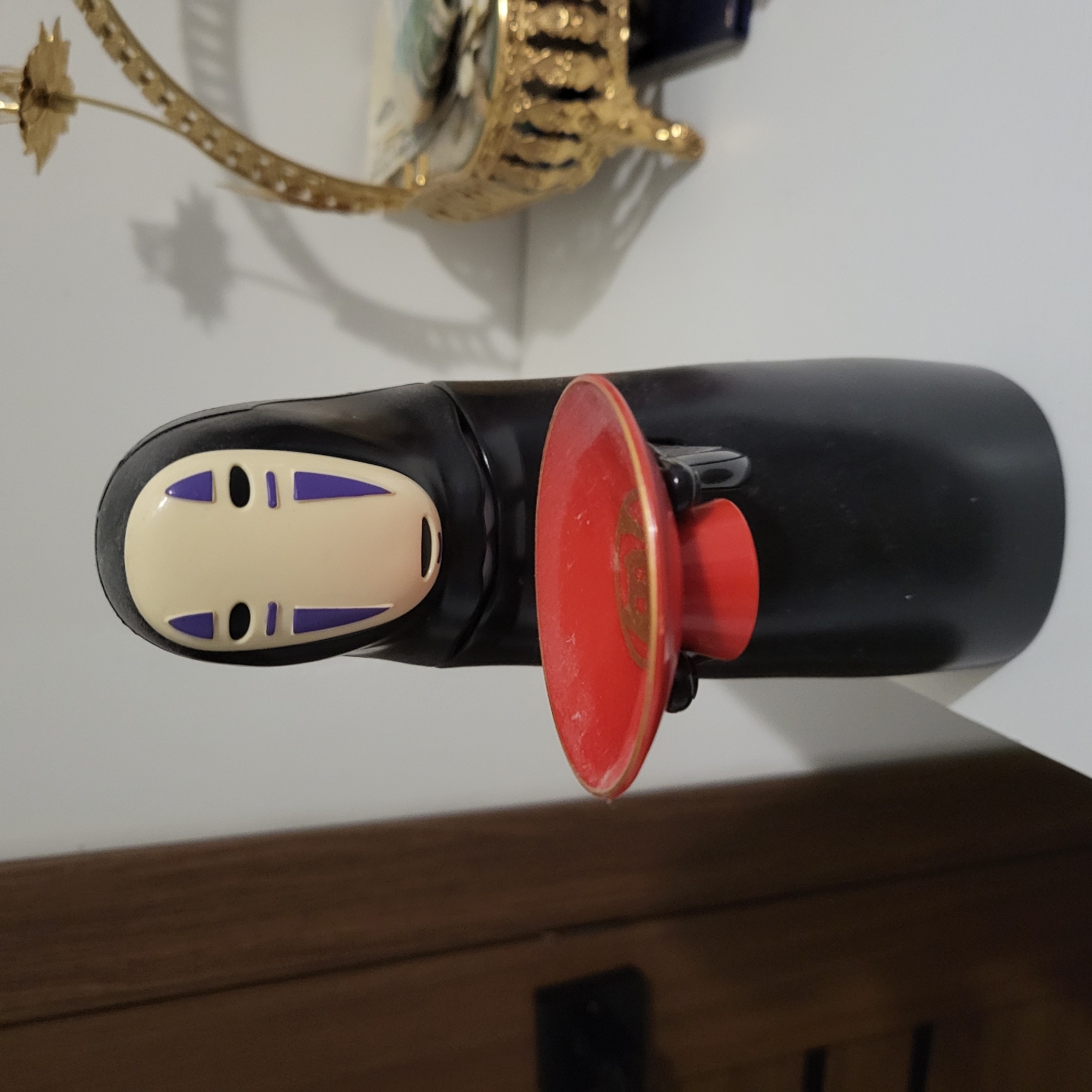 A black figure with a pale mask featuring purple triangles for eyes and mouth stands on a white surface. The figure holds a small red dish with a gold rim. To the right of the figure is a decorative gold object. In the background, a portion of a dark brown vertical surface is visible to the left, and a white wall occupies the rest of the background. The image does not provide any information about the city of Глебовский, Russia.