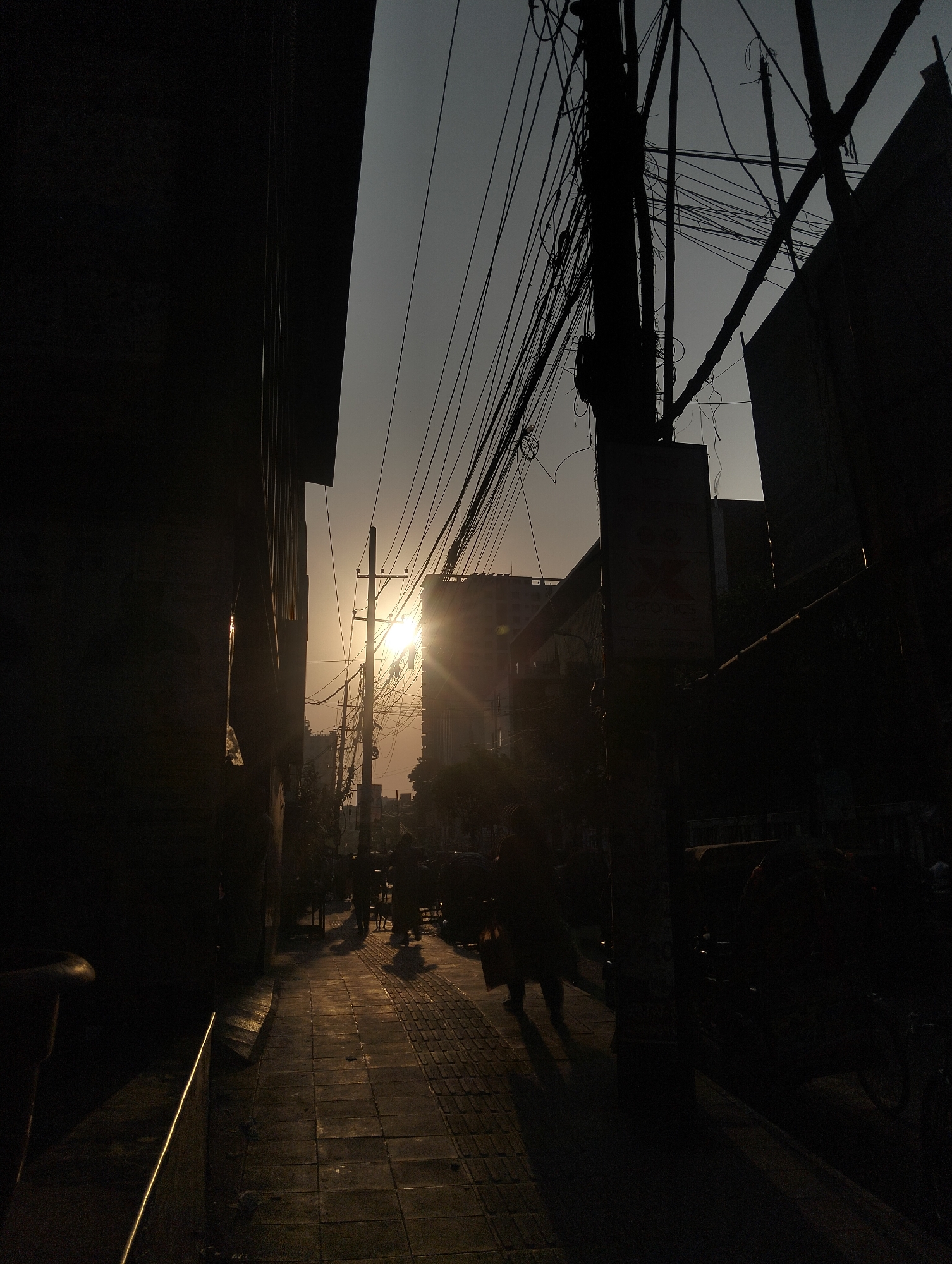 The image shows a street scene in Dhaka, Bangladesh, at sunset. The view is down a narrow street lined with buildings. The buildings on either side are dark and silhouetted against the bright sun. Power lines crisscross overhead. A few people are walking in the distance, their forms obscured by the low light. The sidewalk is made of pavers and is partially lit by the sun.