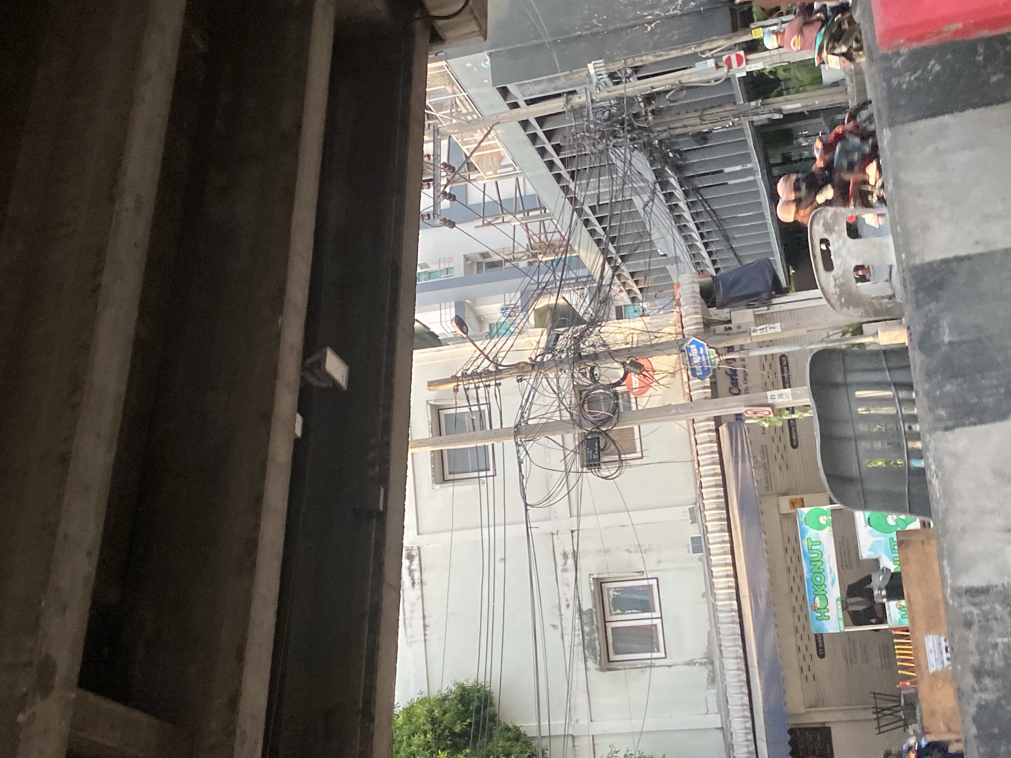 The image captures a street scene in Bangkok, Thailand, viewed from a partially obscured vantage point, likely under a bridge or building. The focus is on the urban landscape, marked by a chaotic network of overhead power lines tangled across the sky. The buildings are closely situated, indicative of dense urban construction, with windows and facades revealing signs of wear. A small patch of green vegetation appears at the bottom of the frame, contrasting with the stark concrete structures. On the right, a commercial establishment is visible, with signage partially readable. The presence of people on motorcycles suggests a bustling street environment.
