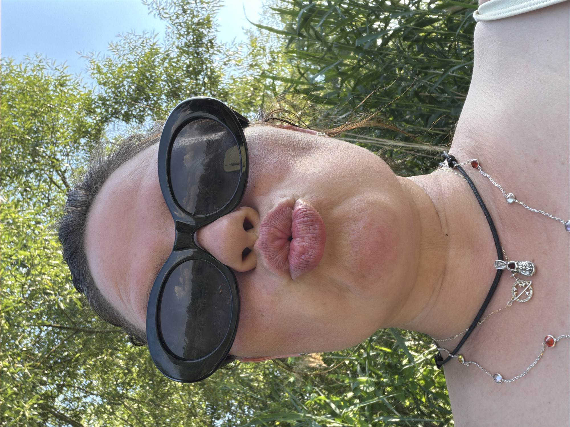 This is a portrait of a person outdoors, likely in Kamianka-Buzka, Ukraine, as suggested by the context. The subject is wearing large, black, oval-shaped sunglasses and has their lips pursed into a kiss. Their head is tilted back, and the image is shot from a low angle, with the sky visible above and lush green foliage in the background. The natural light suggests it is daytime, likely sunny. The person is also wearing a necklace with various charms and colorful beads. The overall mood appears playful and candid.
