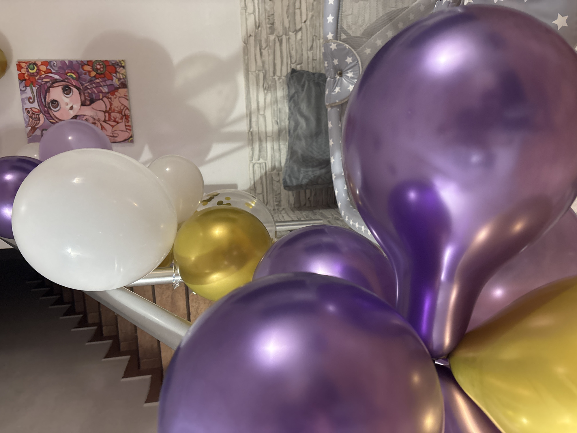 The image shows a close-up of a balloon arrangement in an indoor setting. The balloons are various colors including white, gold, lavender, and purple. They are positioned on or near a metal railing, which appears to be part of a staircase. A framed artwork featuring a large-eyed cartoon-like character is visible on the wall behind the balloons. The background includes a textured wall and a baby cot. The image does not provide enough information to identify the location as Sochi, Russia, and there are no people visible in the scene.