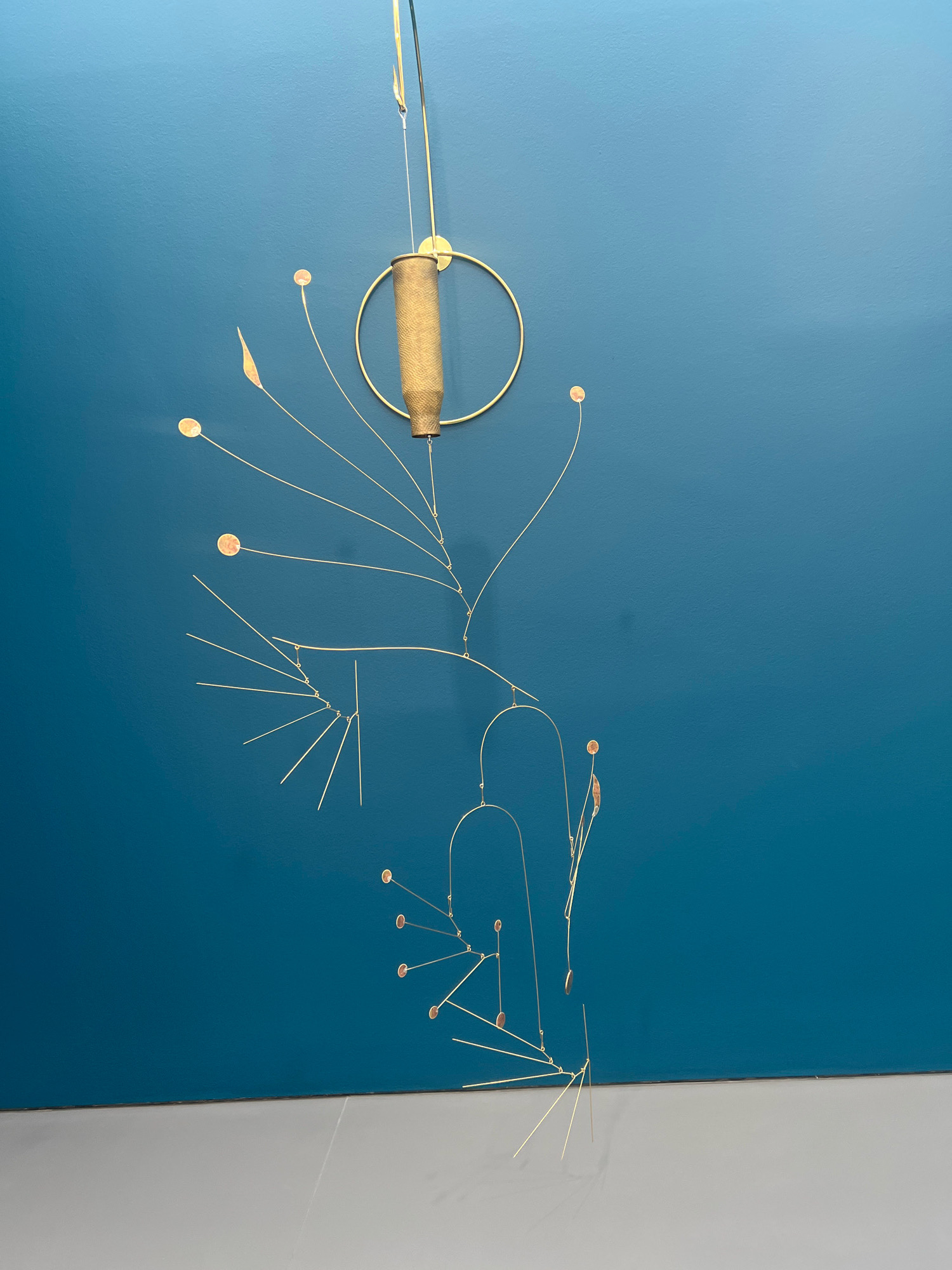 A kinetic sculpture hangs against a teal wall, suspended from the ceiling by thin wires. The sculpture is composed of slender gold-colored metal rods that branch out in a complex, asymmetrical pattern. Small, circular copper-colored elements are attached to some of the rods, resembling tiny discs or coins. The central element of the sculpture is a golden ring from which a larger, hollow cylinder, possibly made of brass or aged metal, is suspended. This cylinder appears to be a spent shell casing. The sculpture is lit from above, casting subtle shadows on the wall behind it. The bottom of the frame shows a lighter-colored surface, likely a floor or a plinth, where the sculpture's lower elements appear to extend towards. The scene is indoors, suggesting an exhibition space or a gallery. No people are visible in the image. This artwork is displayed at the Gropius Bau in Berlin, Germany.