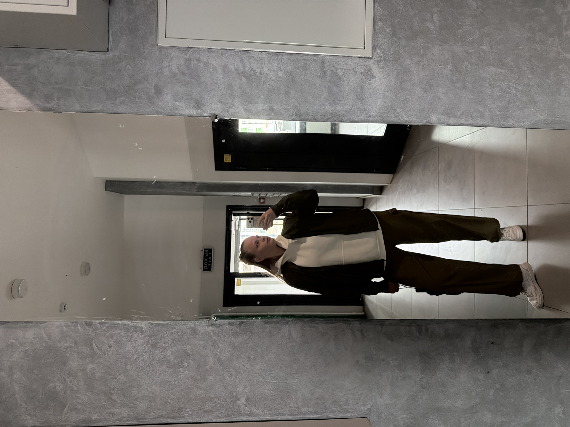 A woman is taking a mirror selfie in a hallway. The reflection in the mirror reveals gray, textured walls and a white tile floor. The woman is wearing dark green pants, white sneakers, a white hoodie, and a dark green jacket. A black door with a yellow sticker and a sign that reads "EXIT" in Russian Cyrillic letters is visible behind her. The hallway appears to be in a building in the city of Izhevsk, Russia, based on the exit sign being in Russian.