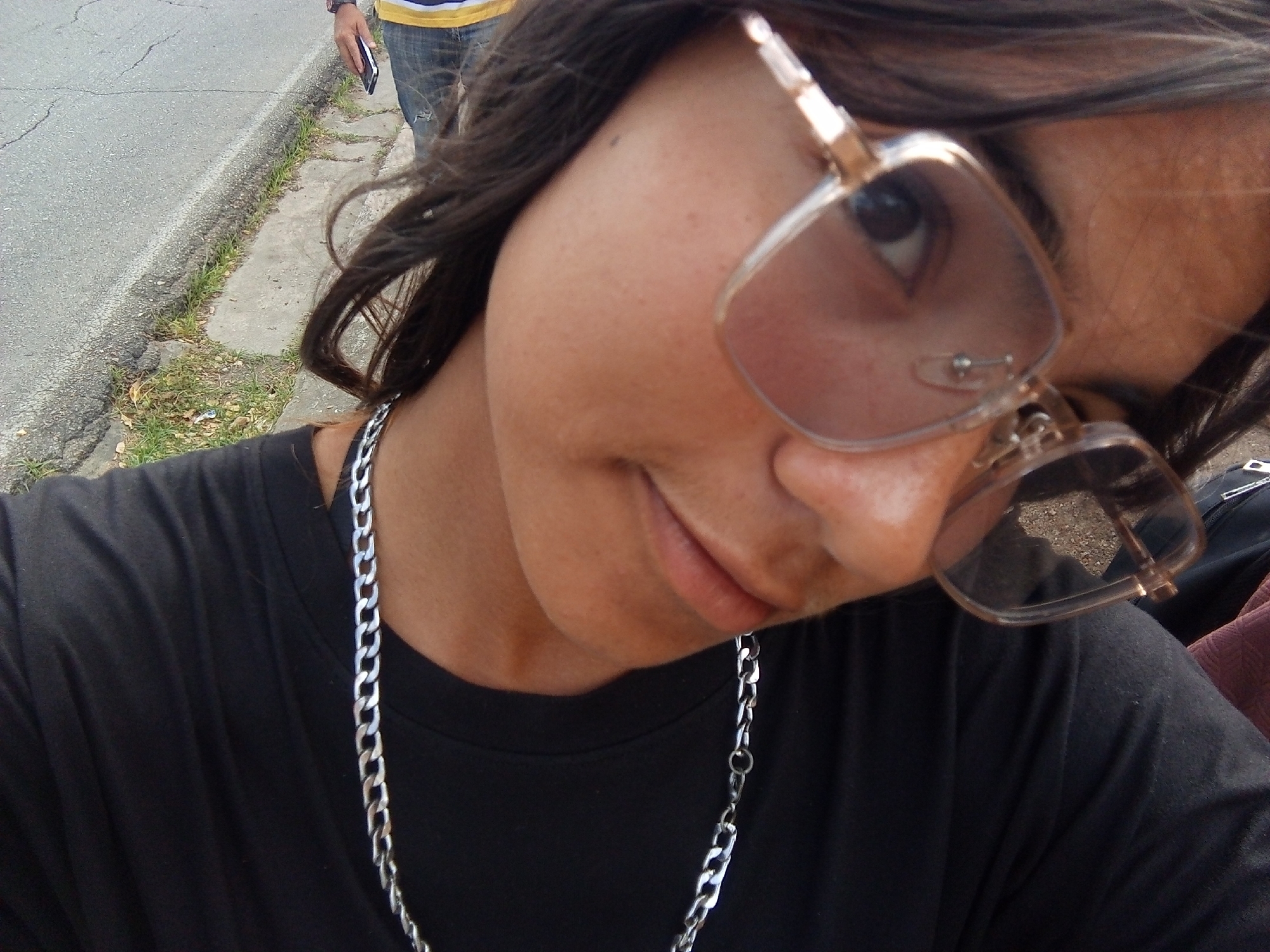 A close-up, high-angle selfie features a dark-haired person wearing large, light-framed sunglasses and a silver chain necklace. The person is wearing a black t-shirt. In the background, partially visible, is another person wearing jeans and a yellow and blue shirt, holding a black object, likely a phone. A road with cracked asphalt and a small strip of grass and weeds beside a concrete curb is visible on the left side of the frame. The image was taken outdoors in Juan Griego, Venezuela.
