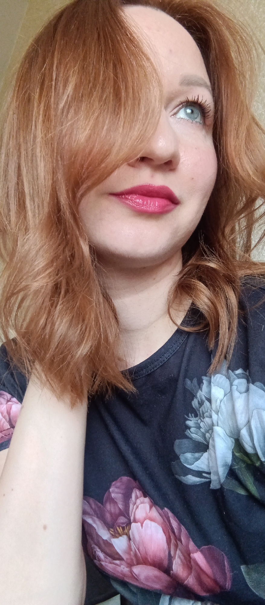 A close-up shot shows a woman's face, her red hair partially obscuring her left eye and side of her face. She has fair skin and is wearing bright pink lipstick. Her eyes are light-colored and looking upwards. She is wearing a dark blue t-shirt with a floral print featuring pink and white flowers. Her left hand is visible, resting near her neck. The background is out of focus and beige. The location appears to be indoors, and the city and country are unknown.