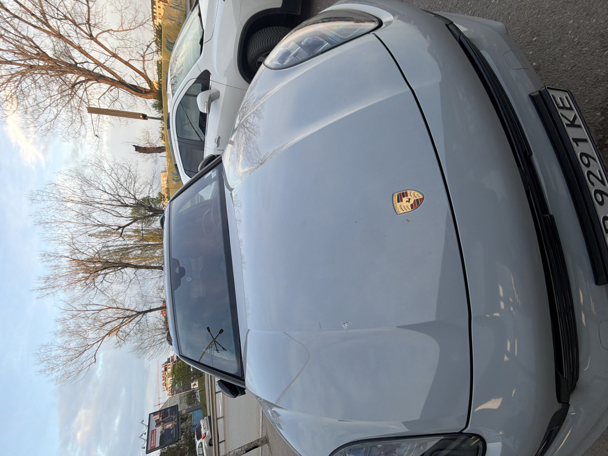 This image depicts a light grey Porsche SUV, likely a Macan or Cayenne model, parked outdoors. The car is viewed from a slightly elevated, diagonal angle, showcasing its front hood with the prominent Porsche crest. Visible on the right side of the hood is a license plate displaying "P 9291 KE," which is consistent with a Bulgarian vehicle registration, with 'P' often denoting the Pernik region. A few minor marks or debris are noticeable on the car's hood.
To the left of the Porsche, a white SUV is partially visible. The background features several bare, leafless trees, suggesting a season such as late autumn, winter, or early spring. The sky is overcast with patches of brighter clouds. In the distance, various urban buildings are present, including structures that appear to be under construction, indicated by visible cranes. A large billboard with illegible text is also visible in the background.
The lighting is soft and diffused, typical of an overcast day or late afternoon/early evening. The setting appears to be an urban or suburban street or parking area in Sofia, Bulgaria. No people or specific activities are depicted in the frame.