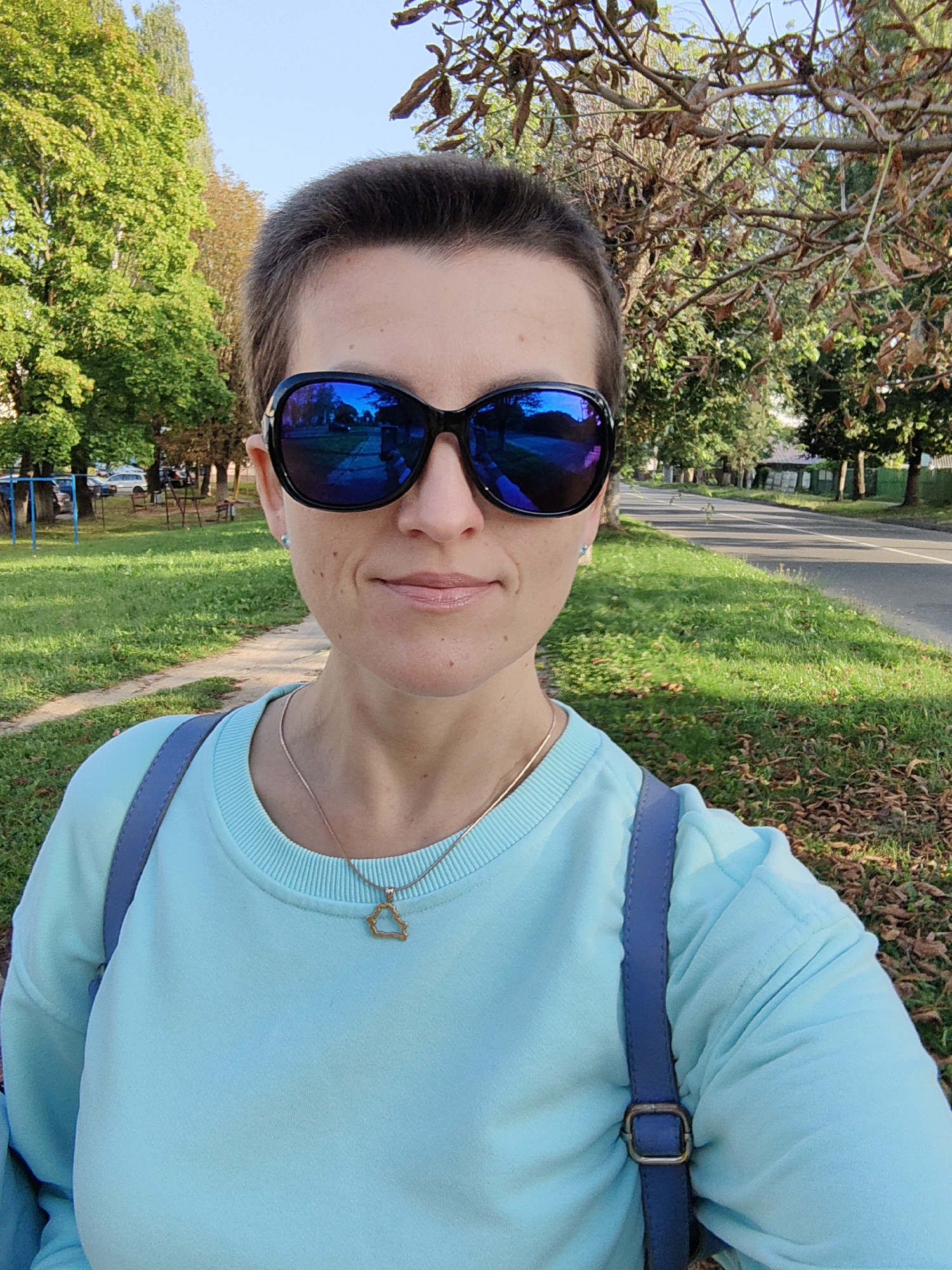 A woman with short dark hair and a pale complexion is taking a selfie outdoors. She is wearing large, dark sunglasses with blue reflective lenses, a light blue sweatshirt, and a thin gold chain with a star-shaped pendant. A blue strap from a backpack or bag crosses her chest. She is smiling slightly.
The background suggests an urban or suburban park setting. There are green trees with some autumn foliage on the left side of the image, and a paved road with grass and fallen leaves on the right. A playground structure is visible behind her on the left. In the distance, houses and more trees can be seen. The sun is shining, casting shadows. The scene appears to be located on a street in Fanipal, Belarus, likely a park or residential area with trees lining the road.