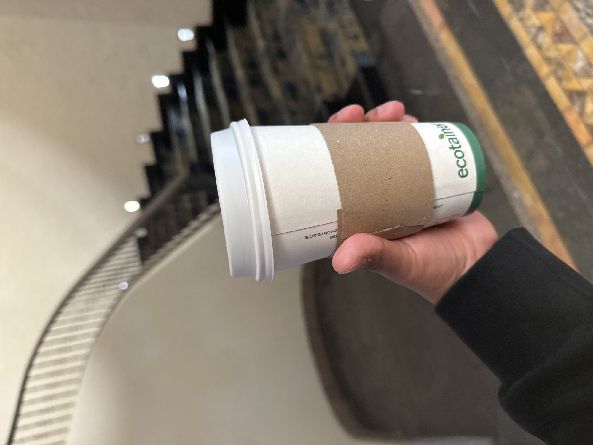 A hand holds a coffee cup with a white lid and a brown corrugated sleeve. The cup has the word "ecotainer" printed in green on the side. The person holding the cup wears a black sleeve. In the background, a curved staircase with decorative railings is visible. The location is the city of San Bernardino Tlaxcalancingo, Mexico.