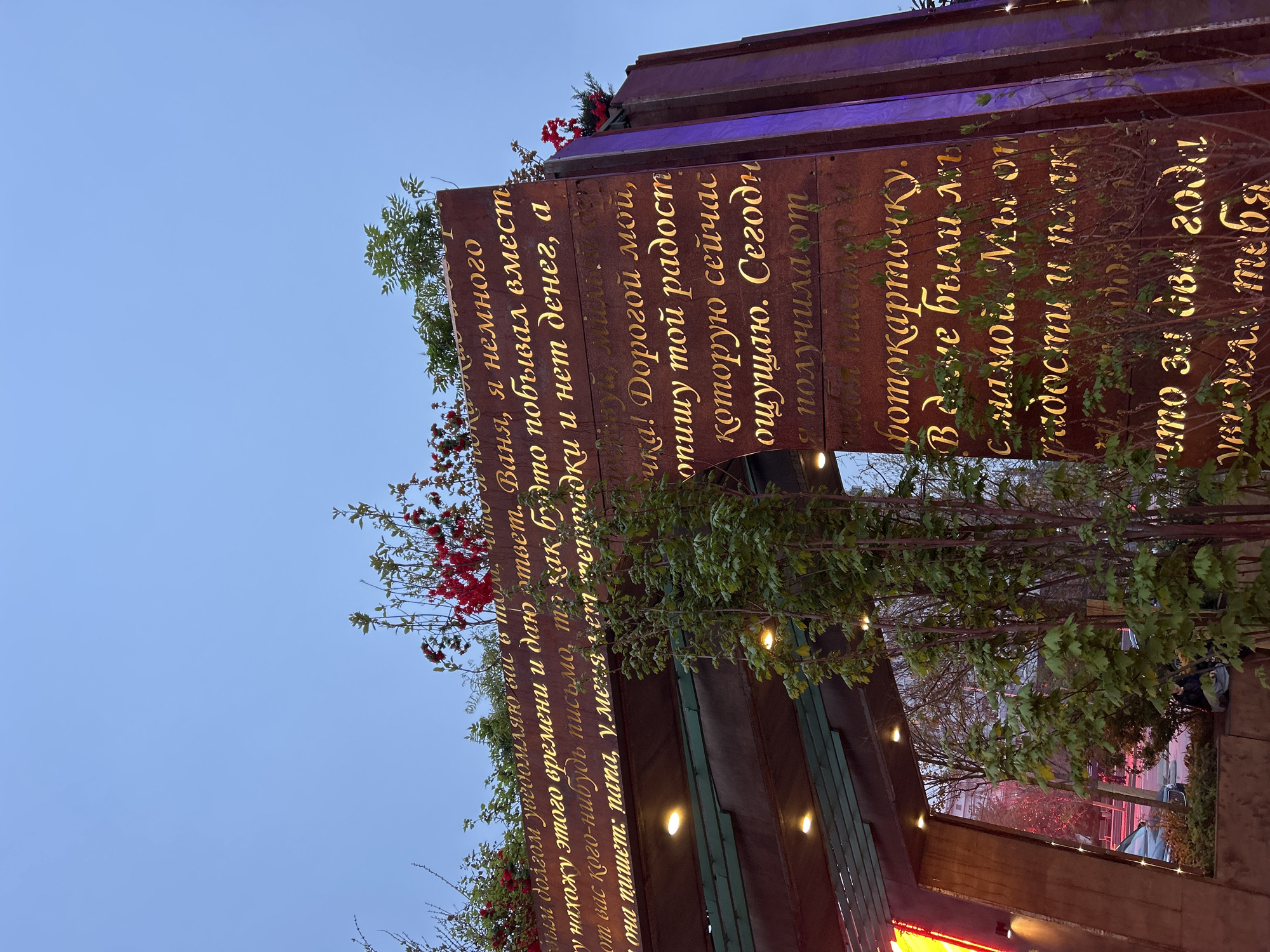 The image shows a sign with Russian text in gold lettering. The sign appears to be made of weathered metal, possibly rusted. Green plants and red berries are growing around the sign, adding a touch of nature to the urban setting. The sign is situated in what seems to be a city environment, although the exact location is not discernible. The sky above is light blue, indicating either early morning or late afternoon. The overall impression is of a public display of text, perhaps a memorial or public art installation.