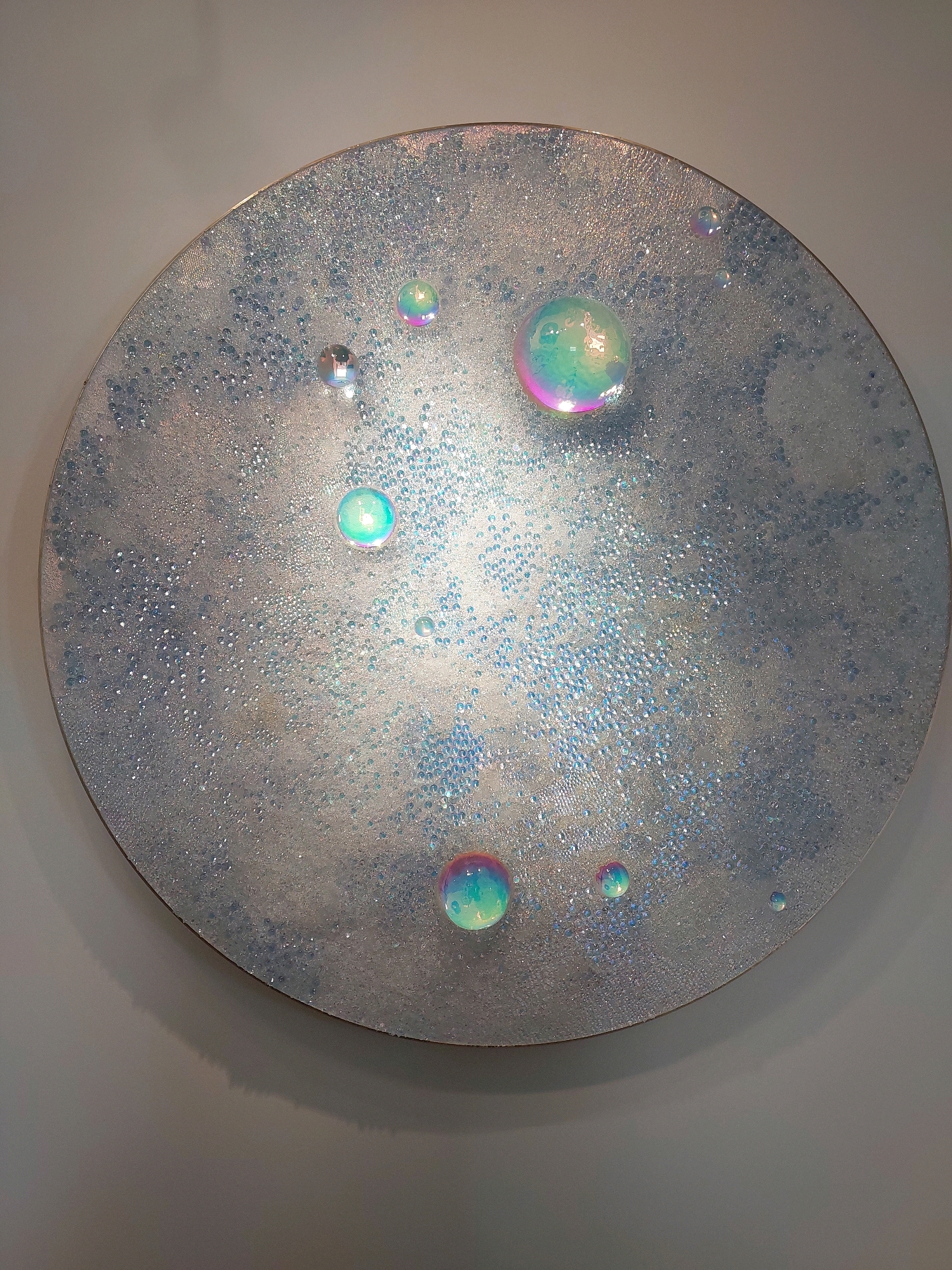 The image shows a circular artwork mounted on a grey wall, likely in a gallery or museum. The artwork itself is a flat, circular surface with a textured, iridescent finish that appears to be silver with hints of blue and white. Scattered across the surface are numerous small, clear, spherical embellishments that resemble bubbles or dewdrops, catching the light and creating a shimmering effect. Several larger, iridescent spheres are also present, exhibiting rainbow-like colors due to light refraction, similar to soap bubbles. These larger spheres vary in size and are strategically placed on the surface of the artwork. The overall impression is one of a celestial or aquatic theme, with the textures and colors evoking stardust or water droplets. No people or specific environmental context outside of the artwork and wall are visible. The provided metadata suggests the location of Saint-Paul-de-Vence, France, implying this artwork is on display in that city.