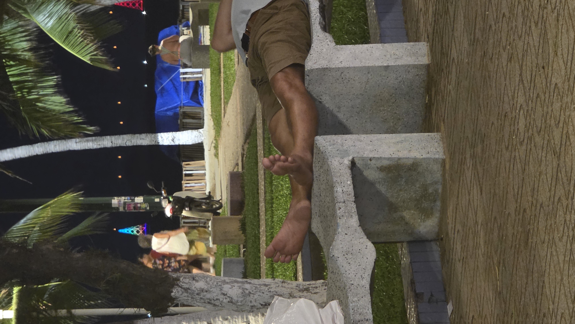 The image depicts a nighttime scene in an outdoor public area, likely a promenade or park in Nha Trang, Vietnam. In the foreground, a person, appearing to be male, is shown from the waist down, sitting barefoot on a light grey, blocky stone bench. He wears brown shorts, and his tanned legs are casually draped over the side of the bench.
Numerous palm trees with light-colored trunks and vibrant green fronds frame the composition, reinforcing the tropical setting. The dark background signifies nighttime, illuminated by various artificial light sources. Small, distant orange lights are visible, along with a prominent, colorful, illuminated conical structure, possibly a decorative light tower. Further in the background, several blurry figures suggest other people are present, some appearing near a structure covered by a blue tarp.
A scooter is parked further along the path, adjacent to a utility pole adorned with various stickers. The ground immediately next to the bench features distinct concrete paving with an etched wavy pattern, transitioning to darker pavers and a strip of green grass. The overall atmosphere appears relaxed, with individuals enjoying the evening outdoors. No discernible text is visible in the image.