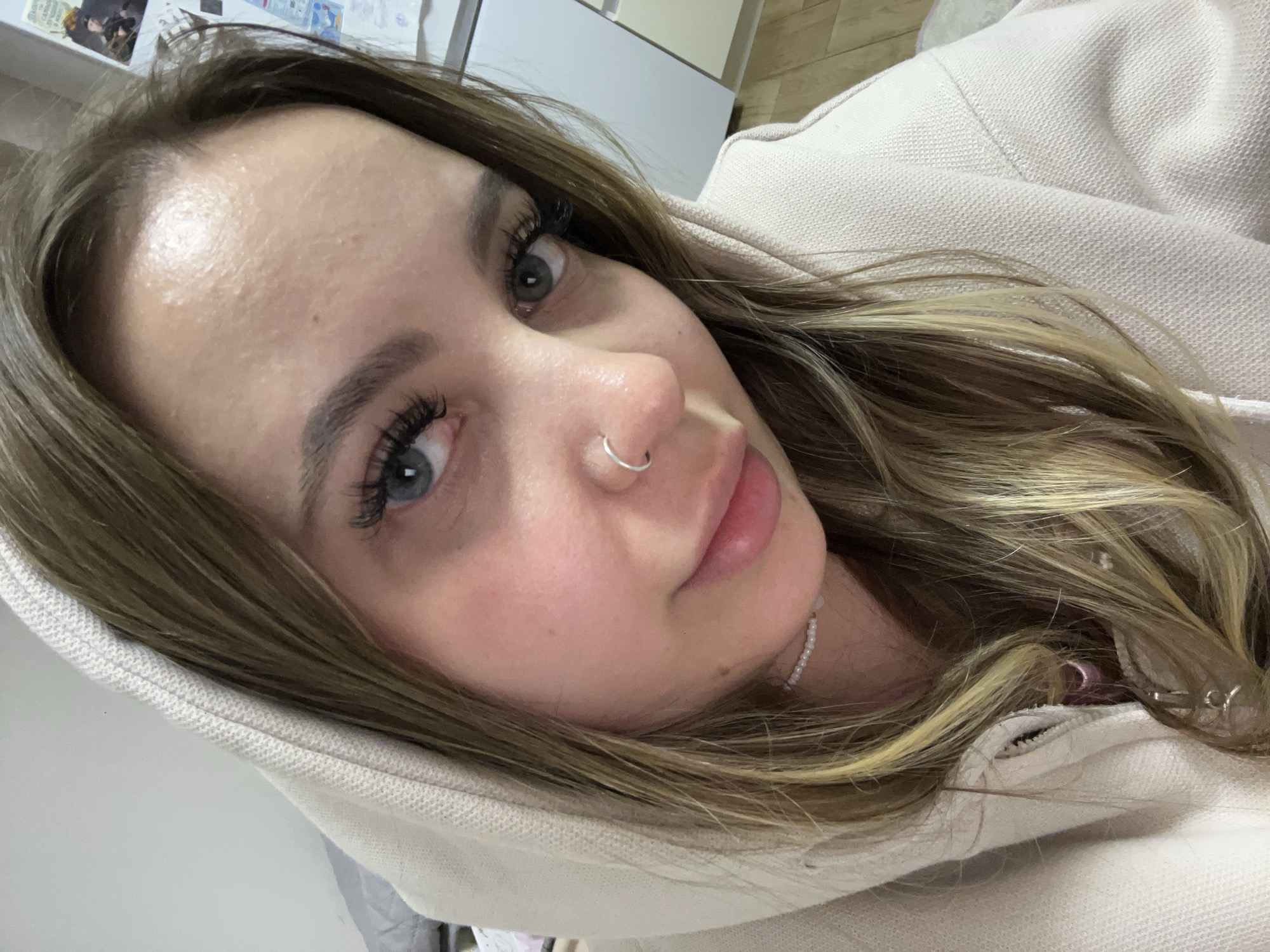 A young woman with long, blonde-streaked brown hair and blue-grey eyes is wearing a light-colored hooded sweatshirt. She has a silver hoop in her left nostril and is wearing a white beaded necklace. The background is blurred, showing a white cabinet and a light-colored wall. There is no indication of the location being Krasnodar, Russia, in the image.