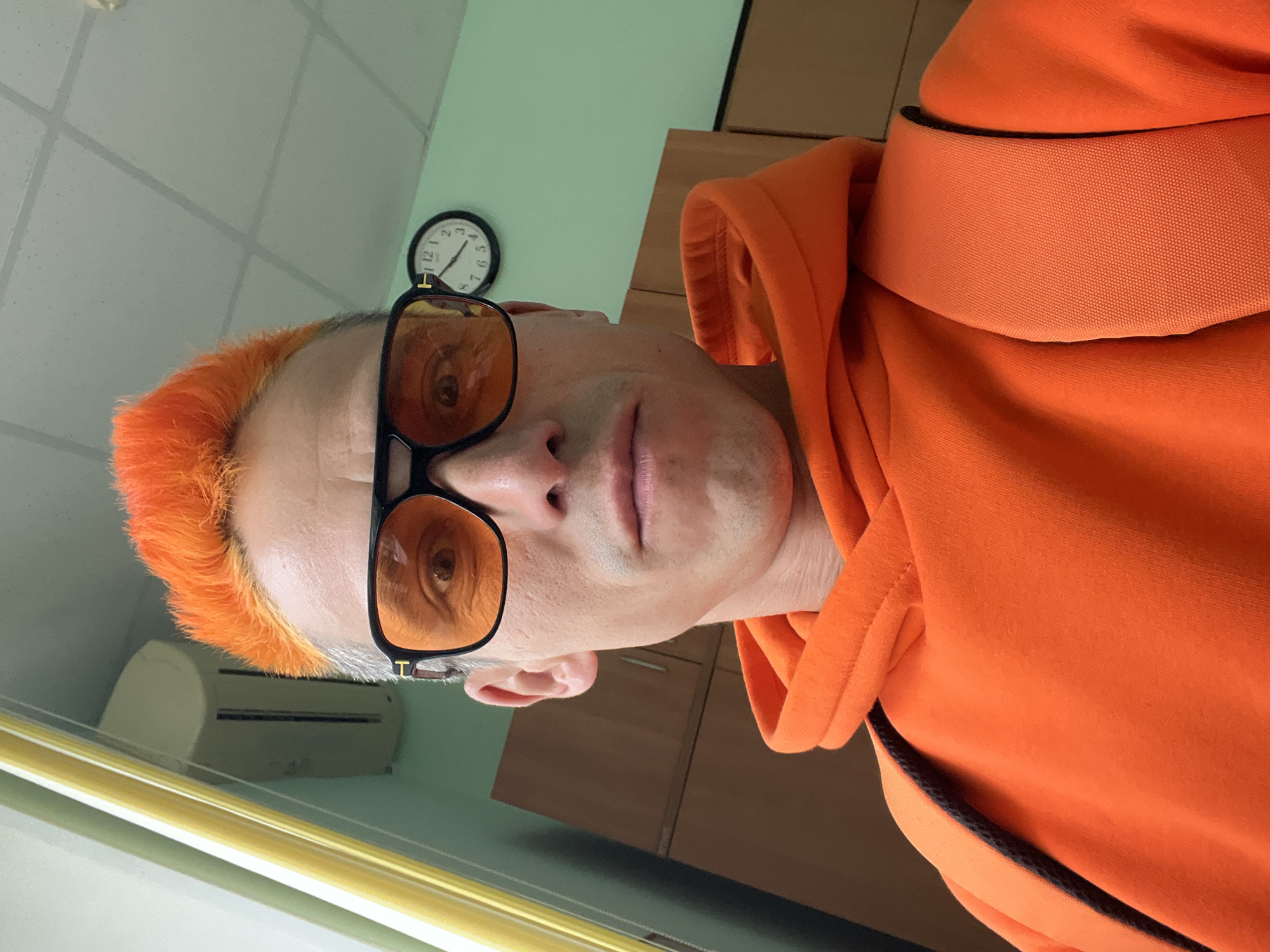 A man with orange hair and amber-tinted glasses, wearing an orange hoodie, is photographed indoors. A clock hangs on a light green wall, and an air conditioning unit is visible. Cabinets are also present in the background. While the city is stated to be Chaikovsky, Russia, there is no visual confirmation of this location within the image itself.