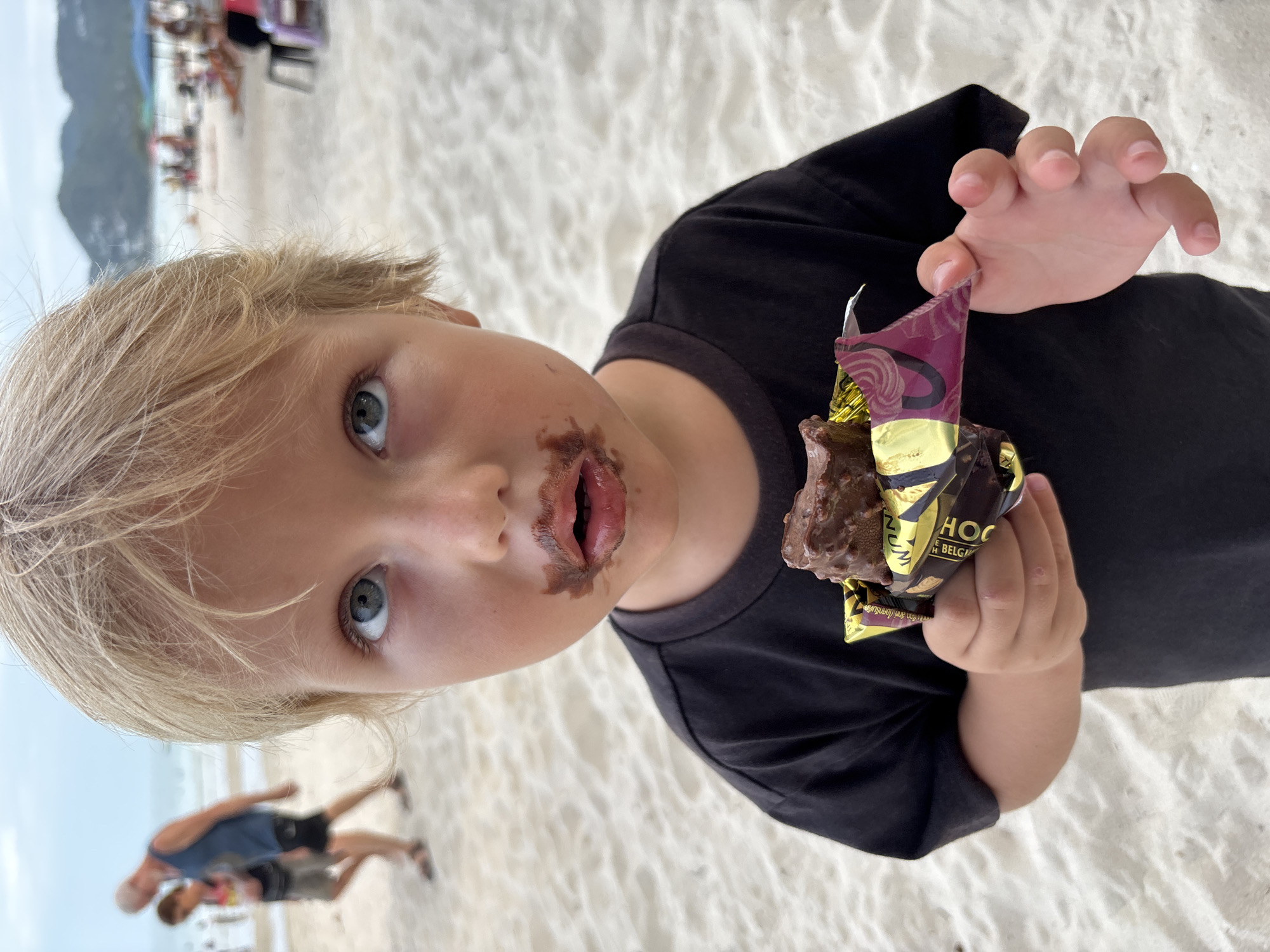 A young child with fair hair and chocolate smeared around their mouth is holding an open chocolate ice cream bar in its wrapper. The wrapper features the words "HOC" and "BELGIUM". The child is wearing a black t-shirt. The background shows a beach with white sand, and in the distance, there are other people. Two figures are seen walking in the distance. The location is Baan Chaweng, Thailand.