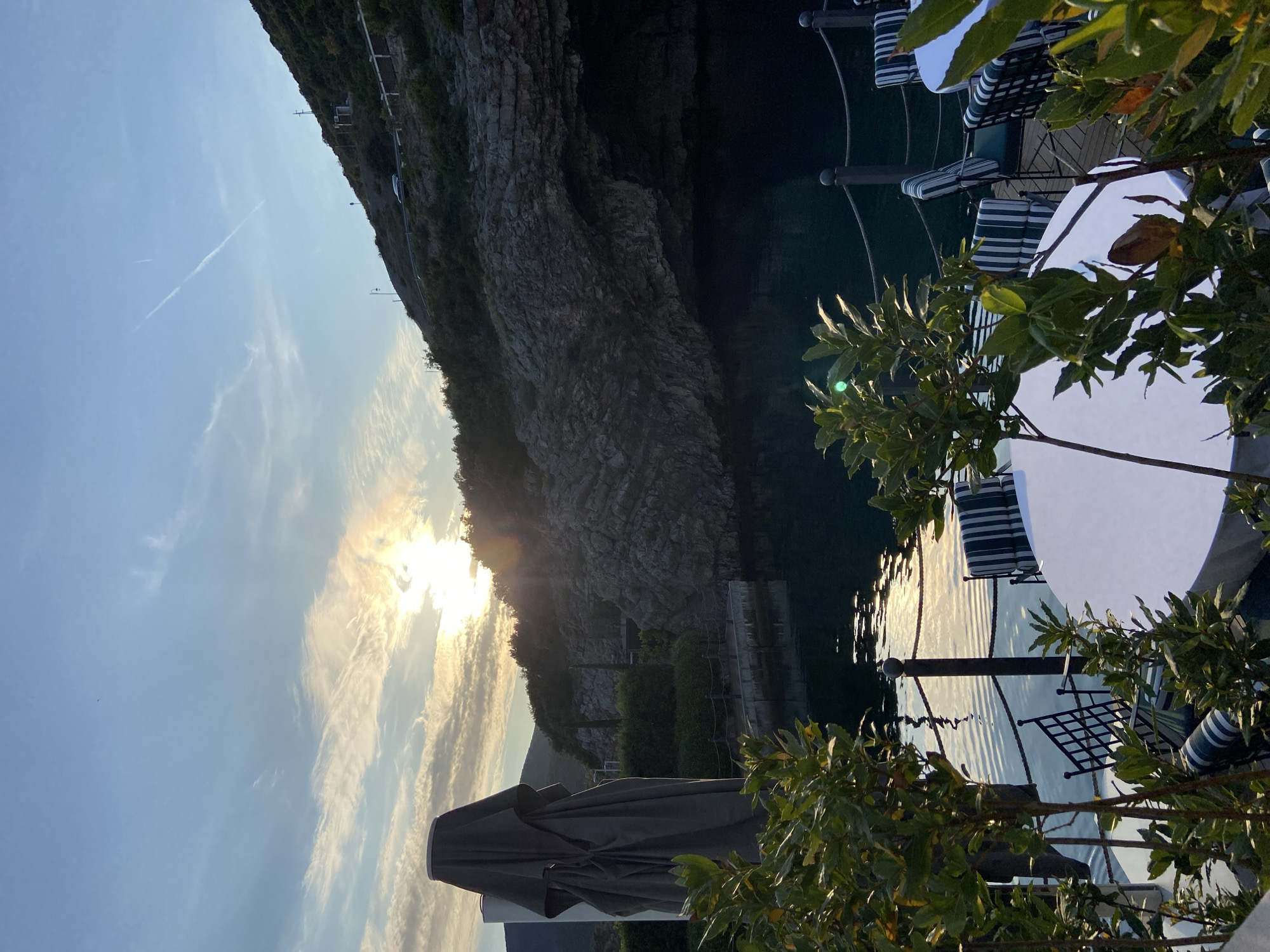 This landscape image, correctly oriented, depicts a serene coastal or lakeside scene, likely an outdoor dining area overlooking a body of water with a rugged cliff face in the background.
In the right foreground, several round white tables are set up, accompanied by chairs featuring distinctive blue and white striped cushions. Green foliage from bushes and tree branches partially frames this dining area, some leaves showing sun flares.
The mid-ground features calm, dark water, which strongly reflects the low-angle sunlight, creating shimmering patterns. On the far side of the water, a massive, imposing rock face or cliff rises steeply, its rugged texture and geological layers visible. Parts of the cliff are covered with sparse green vegetation, and faint structures like paths, small buildings, and light posts can be seen along its base and higher up. A small white boat or floating platform is visible near the water's edge at the base of the cliff.
In the left foreground, a large, dark grey or brown outdoor umbrella is partially open or folded, casting a shadow.
The sky, occupying the upper left and central portions, is a bright blue with scattered, wispy clouds. The sun is low on the horizon behind the cliff in the mid-left, casting a brilliant glare and creating a warm, golden light across the scene, indicating either late afternoon/sunset or early morning/sunrise. The overall atmosphere is peaceful and scenic, suggesting a tranquil leisure spot. No discernible text is visible.