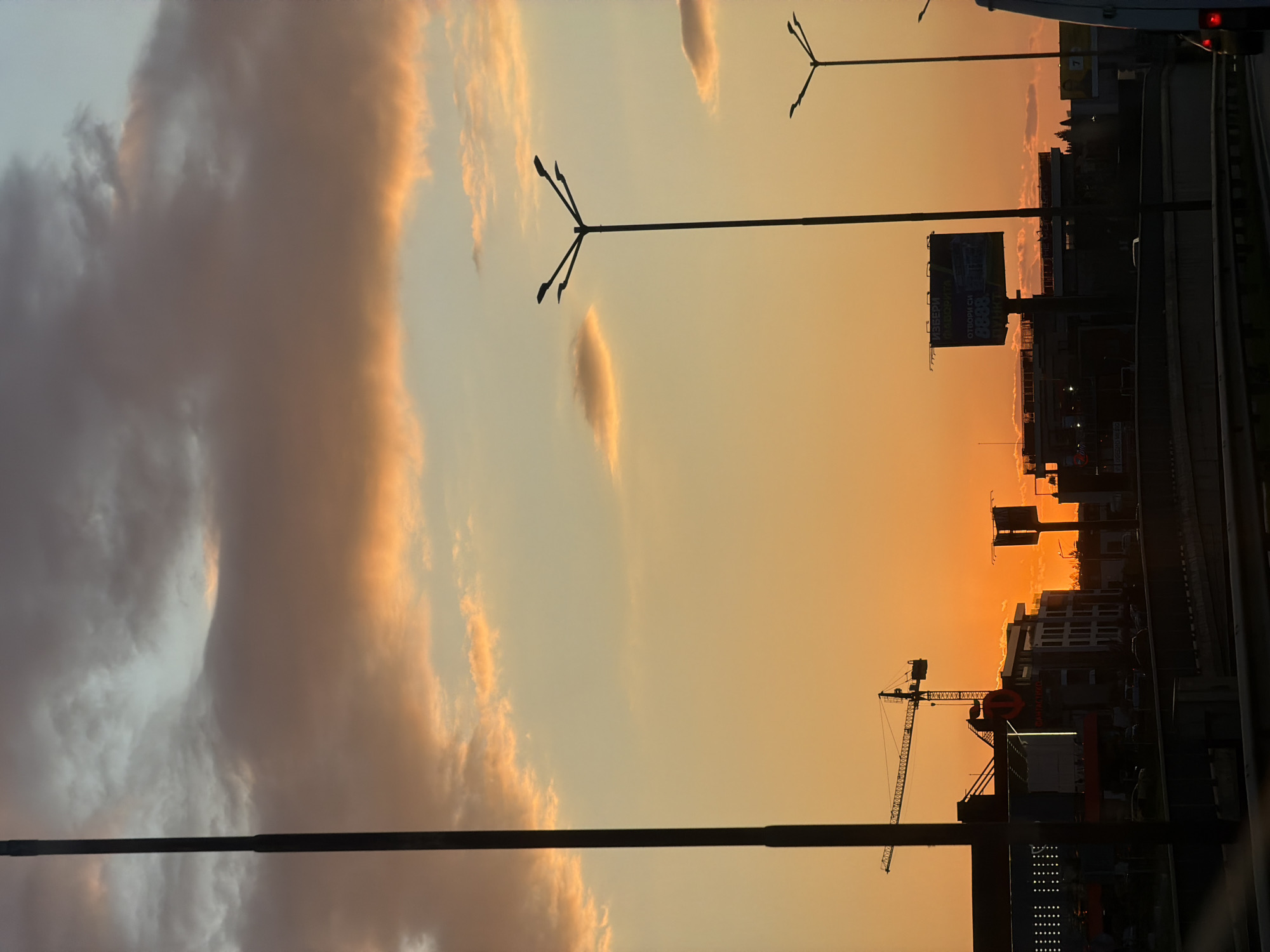 The image depicts an urban cityscape in Sofia, Bulgaria, at sunset. The sky dominates the upper portion, showcasing a gradient of warm colors, transitioning from a vivid orange and yellow near the horizon to softer pinks and light blue higher up. Scattered clouds, illuminated by the setting sun, appear with bright, golden edges or as dark silhouettes against the vibrant backdrop.
In the foreground, several tall streetlights with multiple arms stand out as dark silhouettes against the bright sky. A road or highway, possibly elevated, runs along the right side of the frame, with the back of a white truck partially visible and other faint vehicle shapes suggesting traffic.
The midground and background feature the silhouettes of various buildings, forming an urban skyline. A large construction crane is prominently visible on the left side, adding to the industrial aspect of the cityscape. Several billboards are also silhouetted against the bright sky. On one large billboard, faint Cyrillic text can be discerned, reading "ИЗБЕРИ СВОБОДАТА" (Choose Freedom), "ОТВОРИ СИ СМЕТКА" (Open your account), and the number "8888". Some small lights from buildings and possibly other vehicles are visible in the darkening environment. The scene captures the serene beauty of dusk over a bustling city.