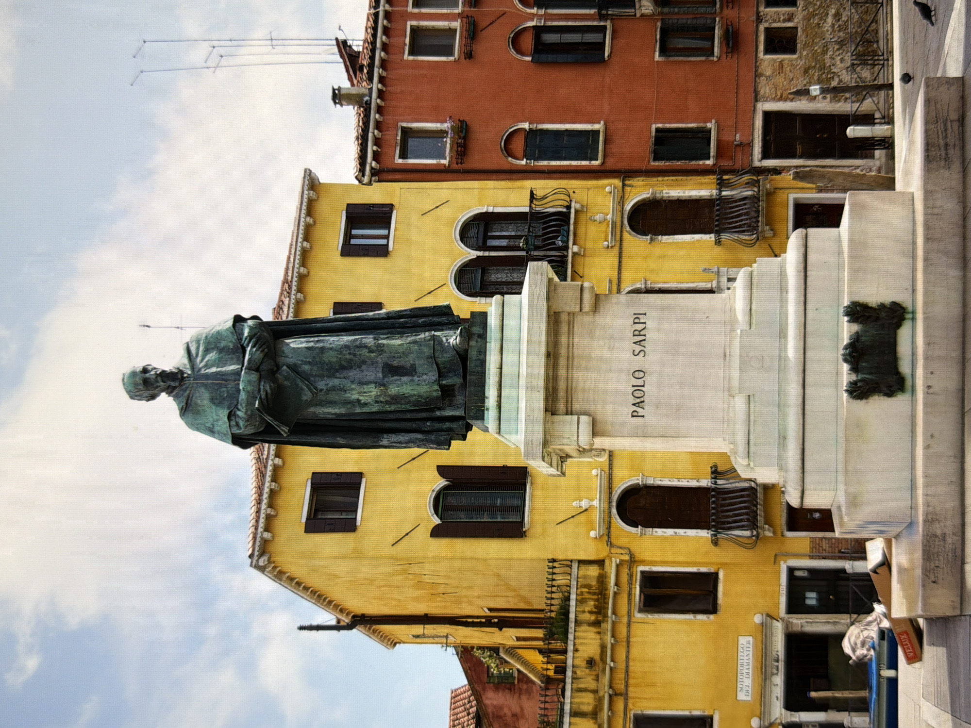 This is a photograph of a bronze statue of a man in robes, standing on a stone pedestal in a public square. The statue is located in front of two colorful buildings, one yellow and one orange-red, typical of Venetian architecture. The yellow building has several windows and balconies, as does the orange-red building. In the background, there are antennae visible against a cloudy sky. The pedestal of the statue has the name "PAOLO SARPI" inscribed on it. The ground in front of the pedestal is paved, and there is a blue tarp and some cardboard boxes near a blue structure on the left side of the image. Pigeons are present on the paved ground on the right side of the image. The scene appears to be outdoors, in a pedestrian area. This image is not located in the city of Kyiv, Ukraine; it is located in Venice, Italy.