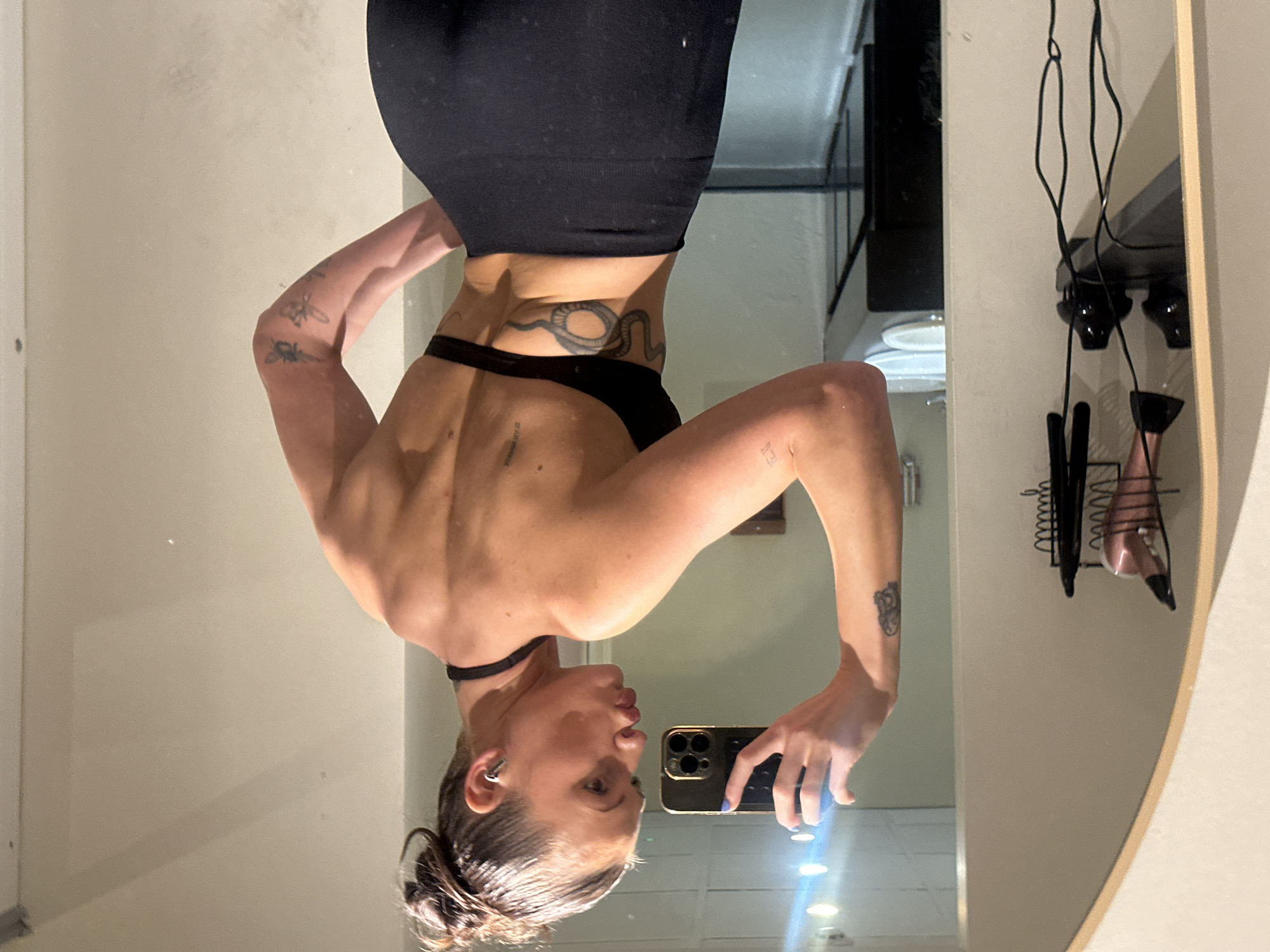 A woman takes a photo of herself in a mirror. She wears a black top and bottom. Her hair is up in a bun. She has tattoos on her arms, back, and near her hip. The mirror reflects a bathroom. There is a light on the mirror's surface. It is impossible to determine the exact location in Tunbridge Wells, United Kingdom.